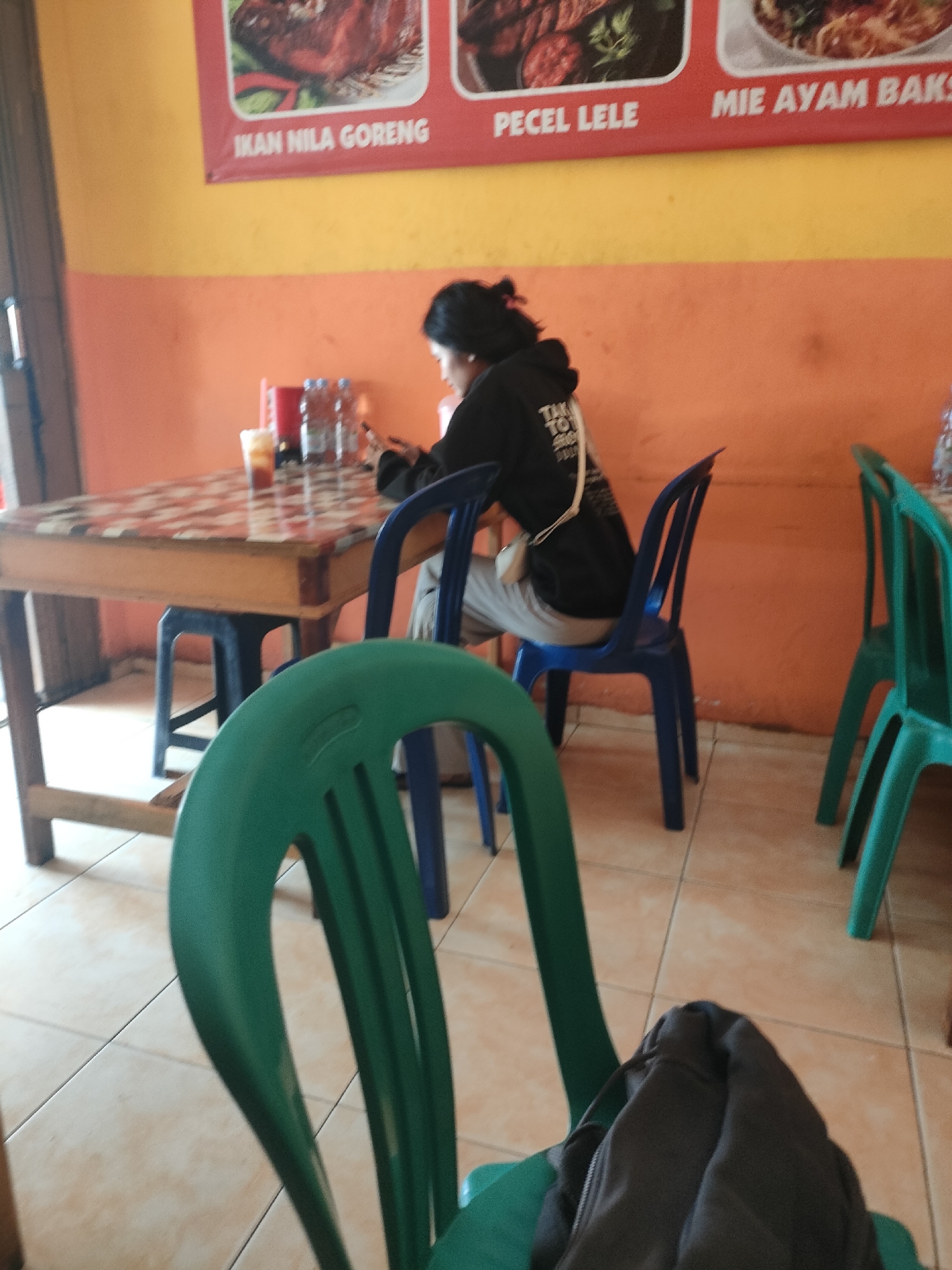 The image shows an indoor scene, likely a restaurant, with a woman sitting at a table, looking down at her phone. She is wearing a black hoodie and light-colored pants. In front of her is a table with a checkered tablecloth, holding several bottles of drinks and a small dish. Two blue chairs are visible, one occupied by the woman and another to her right. In the foreground, a portion of a green chair is visible. The walls are painted in orange and yellow. A banner with text in Indonesian, featuring images of food, is displayed on the upper part of the wall. The flooring appears to be tiled. The scene is likely in a modest, casual eatery, potentially a local food stall or restaurant, common in cities like Pringsewu, Indonesia, where such establishments are prevalent.