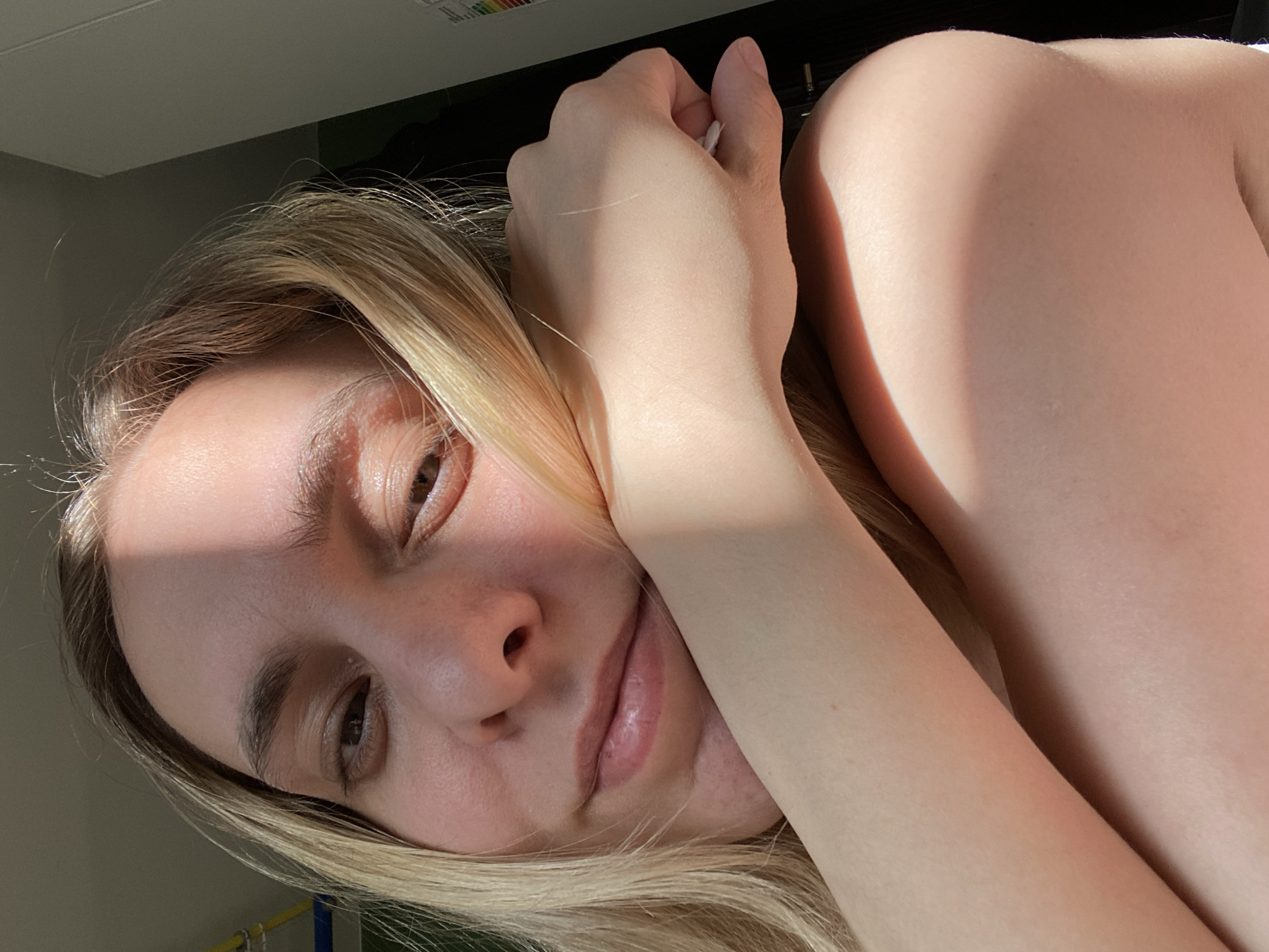 A young woman with blonde hair is looking at the camera. She has brown eyes. She's resting her face on her hands. The light appears to be coming from a window, possibly from outside. The image location is unknown, and it's not possible to confirm the location in Chaikovsky, Russia.