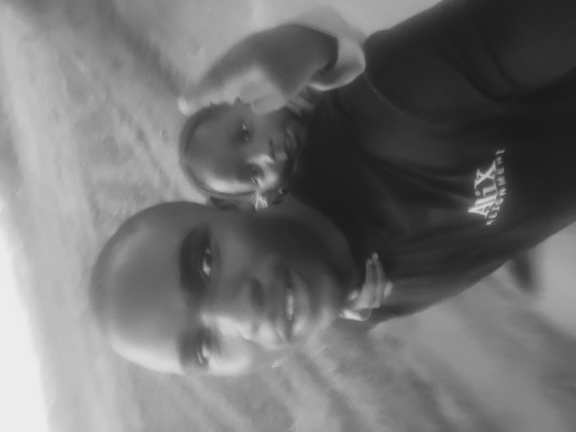 A black and white, slightly blurry, vertical-oriented photograph captures a moment between two individuals, likely in Bukuru, Nigeria. The image is taken at an angle, making it appear tilted.
The foreground features the upper body and face of an adult, possibly male, with a broad smile. The person's eyes are looking towards the viewer. They are wearing a dark-colored top.
Behind and slightly above the adult, a child is visible, perched on their back, suggesting a piggyback ride or similar playful interaction. The child also appears to be looking towards the camera, though their expression is less clear due to blur and angle. The child is wearing a lighter-colored top or hood.
On the adult's dark top, text is visible, though partially obscured and rotated. The word "Alix" is clearly discernible, followed by another word that appears to be "CONIMENT" or something similar, suggesting a brand name or an organization.
The background is indistinct, appearing to be an outdoor setting, possibly a path or ground, given the texture and lack of clear structures. The lighting suggests it might be daytime, but the black and white nature of the image and the blur make it difficult to determine specific weather conditions or exact time of day. The overall emotion conveyed by the adult's smile is one of happiness or joy.