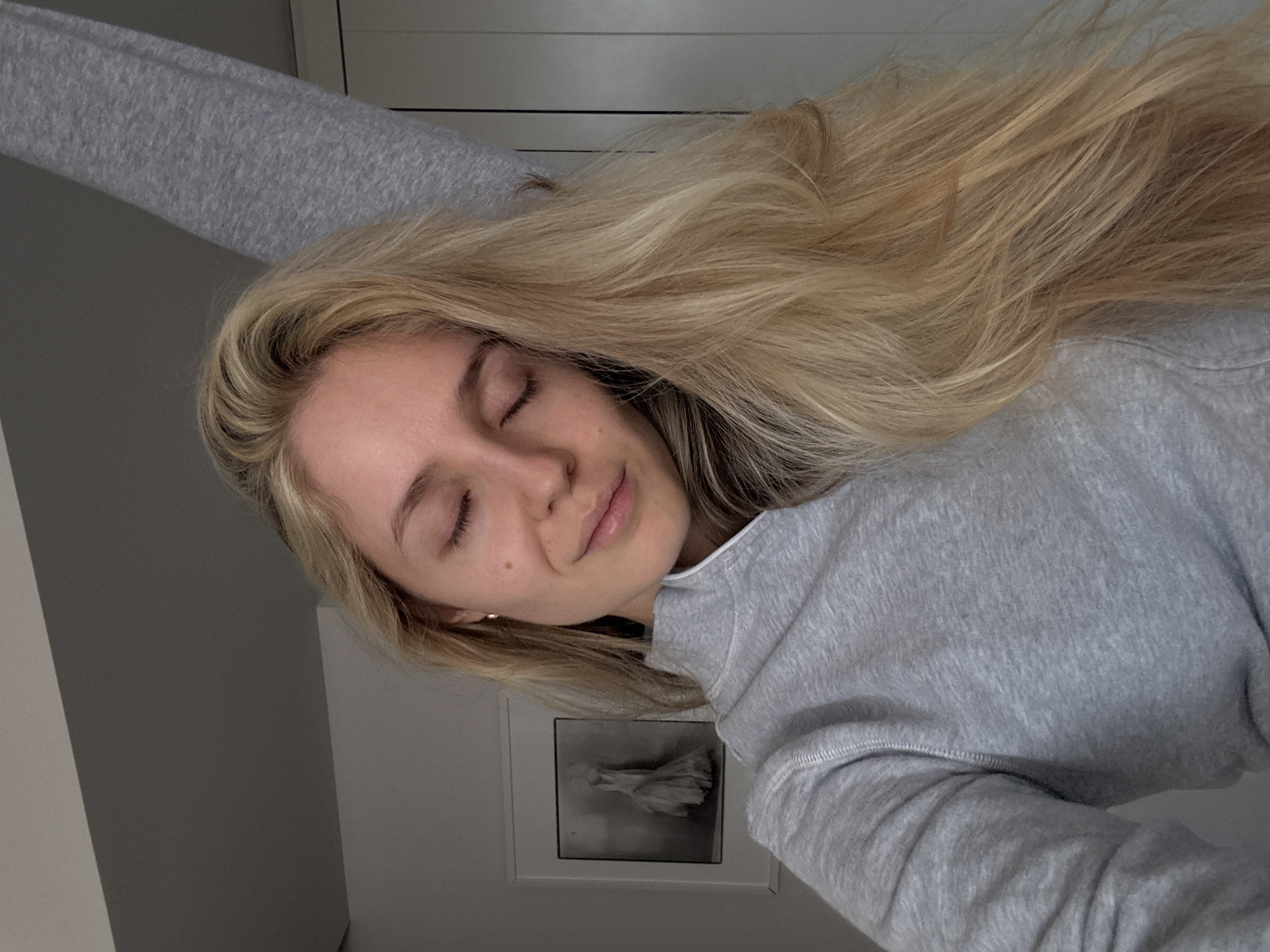 A fair-skinned young woman with long blonde hair rests, eyes closed, wearing a gray sweatshirt. The background is blurred, showing light gray walls. A framed picture hangs on the wall behind her. The location is a room in Mexico City, Mexico.