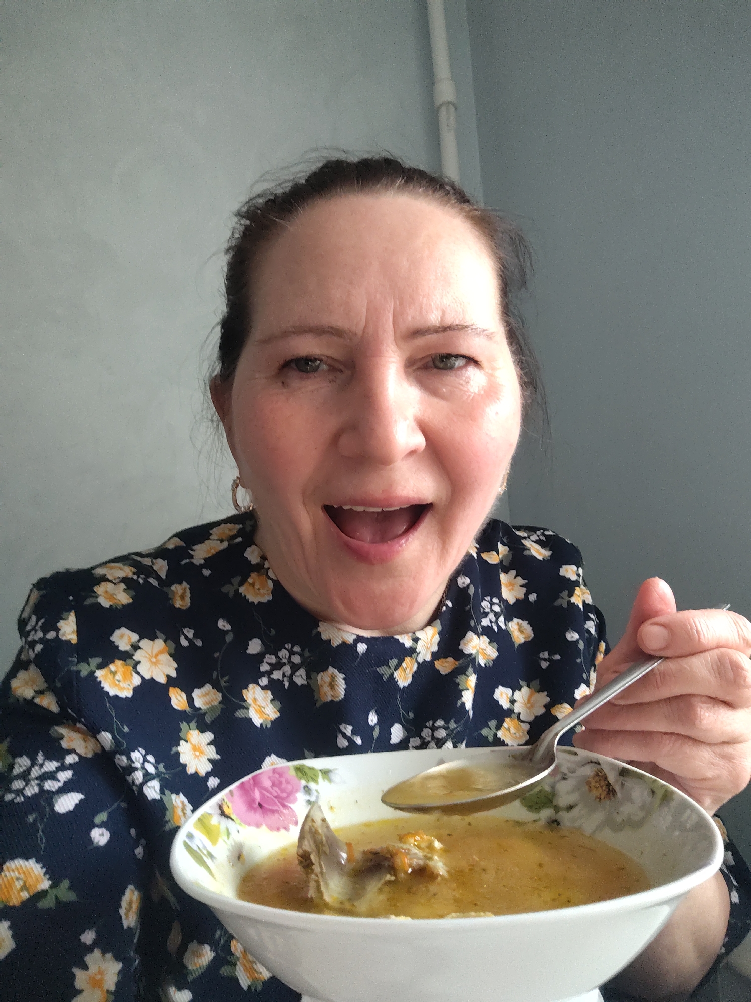 A middle-aged woman with fair skin and brown hair pulled back into a bun is holding a bowl of soup and a spoon. She is wearing a dark blue dress with yellow and white floral patterns. She has gold hoop earrings and her mouth is open, appearing as if she is about to eat the soup. The bowl is white with a floral design, and the soup appears to contain meat and vegetables. The background is a plain, light gray wall. The photo was likely taken indoors. There is no indication that this picture was taken in the city of Sarapul, Russia.
