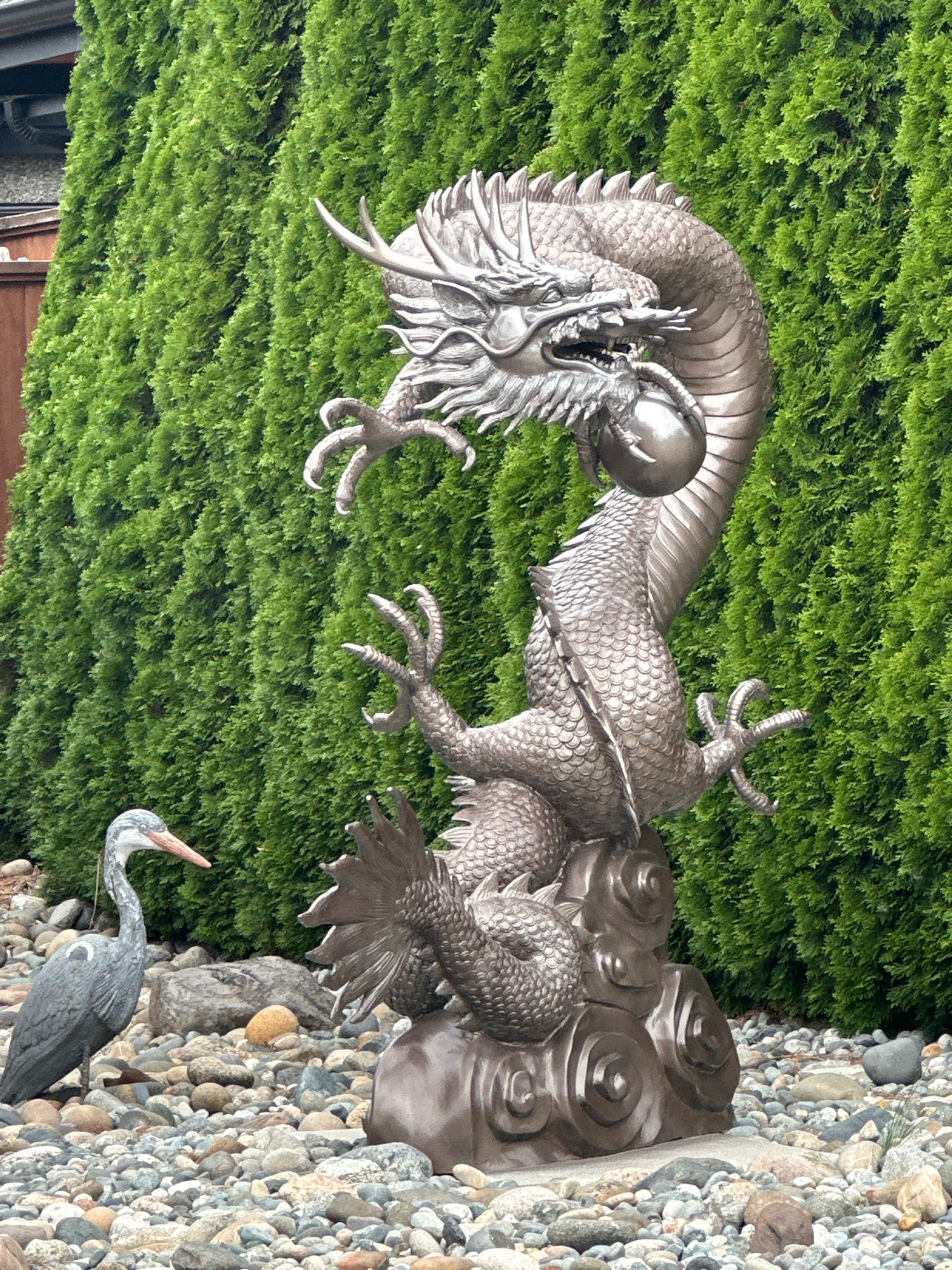 The image depicts an outdoor scene with a statue of a dragon holding a pearl, positioned in front of a dense hedge of green foliage. To the left of the dragon statue, there is a grey heron statue. The ground is covered in various-sized rocks and pebbles. The dragon statue is intricately detailed, showcasing scales, claws, and facial features. The heron statue is positioned lower than the dragon, looking towards it. The background consists of the uniform green hedge and a hint of a building structure on the far left. The scene appears to be a garden or an outdoor decorative display. This image does not contain any discernible information that would allow for its location to be identified within the city of Burnaby, Canada.