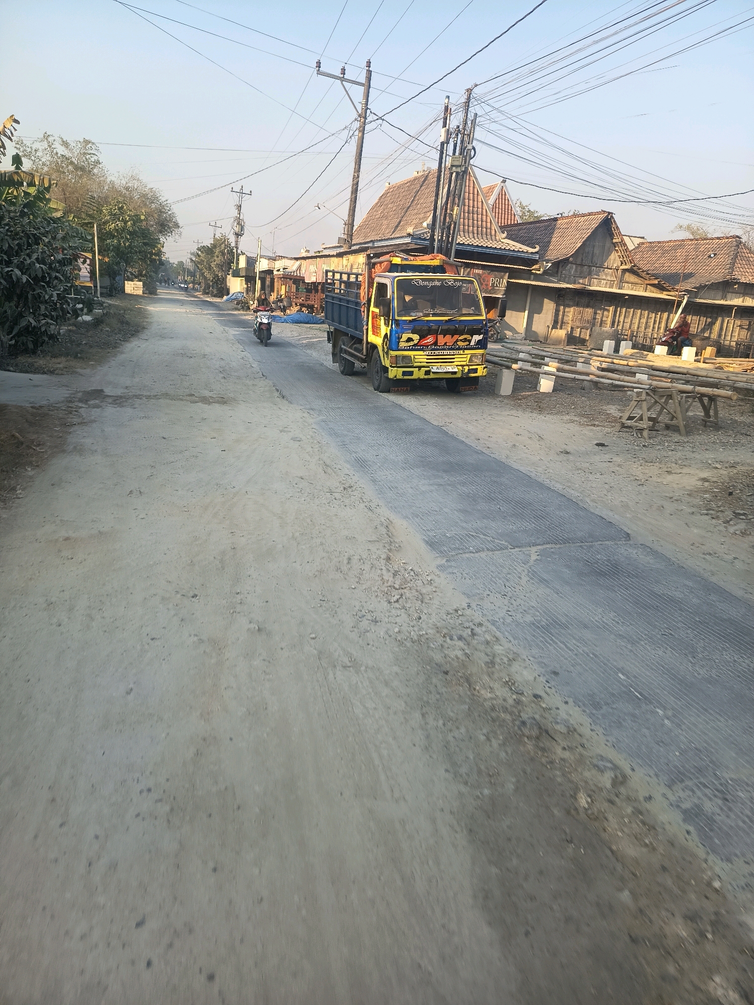 The image depicts a street scene in Pulokulon, Indonesia, characterized by a dusty, unpaved road alongside sections of new concrete pavement. A yellow and blue truck with text that appears to read "POWER" and "Bahan Bangunan" (Building Materials) is parked on the side of the road. In the distance, a motorcycle is being ridden down the street, and several buildings with tiled roofs are visible. The environment appears to be a semi-rural or developing urban area, with visible power lines overhead. There are also construction materials, including bamboo poles, stacked along the side of the road, indicating ongoing development or repair work. The sky is clear and a light blue, suggesting daytime with mild weather conditions.