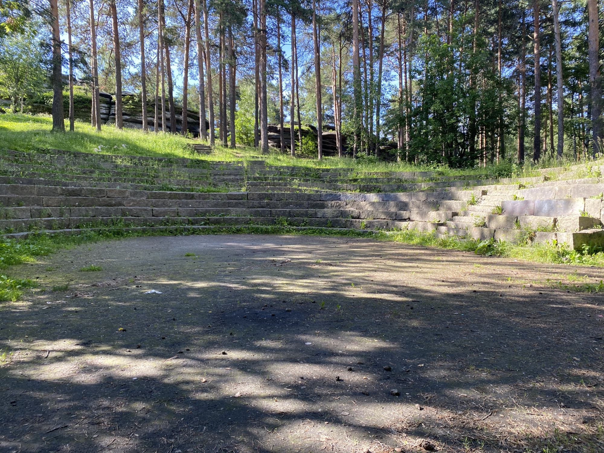 The image shows an outdoor amphitheater made of stone benches surrounded by trees. The amphitheater is in a park-like setting, with grass and trees visible in the background. The ground in front of the amphitheater is covered with dirt and fallen leaves. The lighting suggests it is daytime. This appears to be an abandoned or disused structure, with some vegetation growing between the stone seats. The location is identified as an amphitheater in Yekaterinburg, Russia.