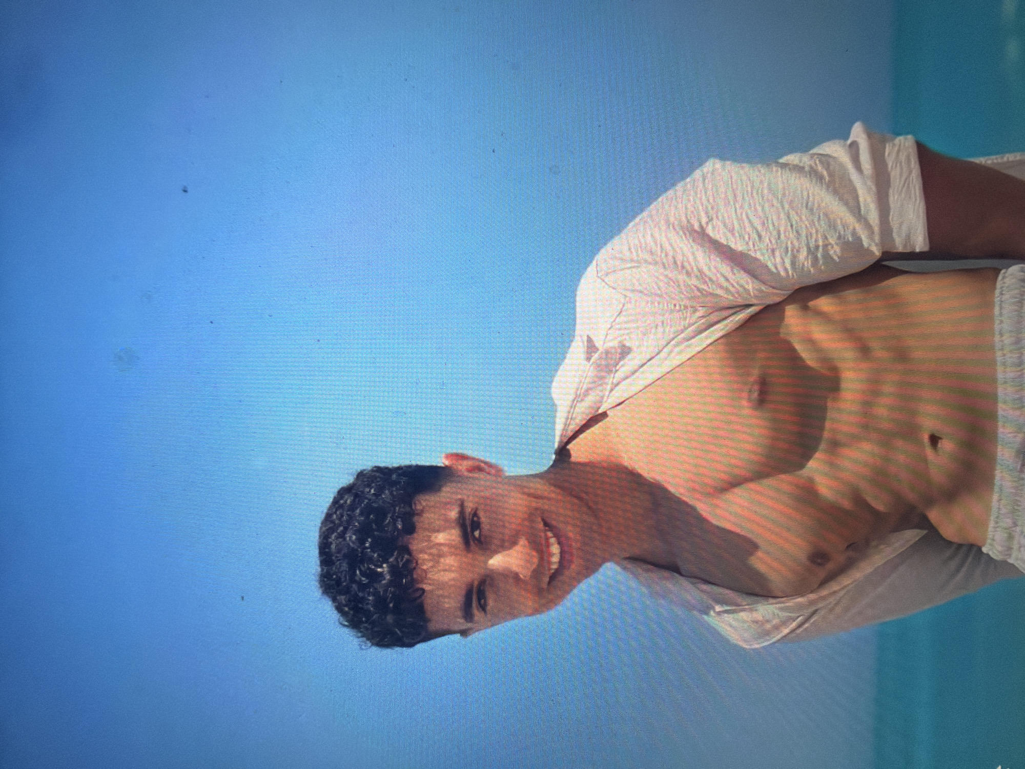 The scene, set in the city of Shumen, Bulgaria, features a young adult male captured from the waist up in a coastal or beach environment. The subject has dark, curly hair and is shown smiling broadly with teeth visible, gazing directly towards the viewer. He is positioned slightly to the viewer's left. He wears an unbuttoned, light-colored, long-sleeved shirt, which hangs open to reveal his muscular chest and well-defined abdominal muscles. Light-colored shorts or swim trunks are visible at the bottom edge of the frame. The background consists of a clear, bright blue sky transitioning into a body of turquoise water, indicative of an ocean or sea. The entire image displays a distinct pixel grid pattern, consistent with a photograph taken directly from a digital screen. Multiple small, dark specks, likely dust or imperfections on the screen, are scattered across the background, particularly in the upper sky portion.