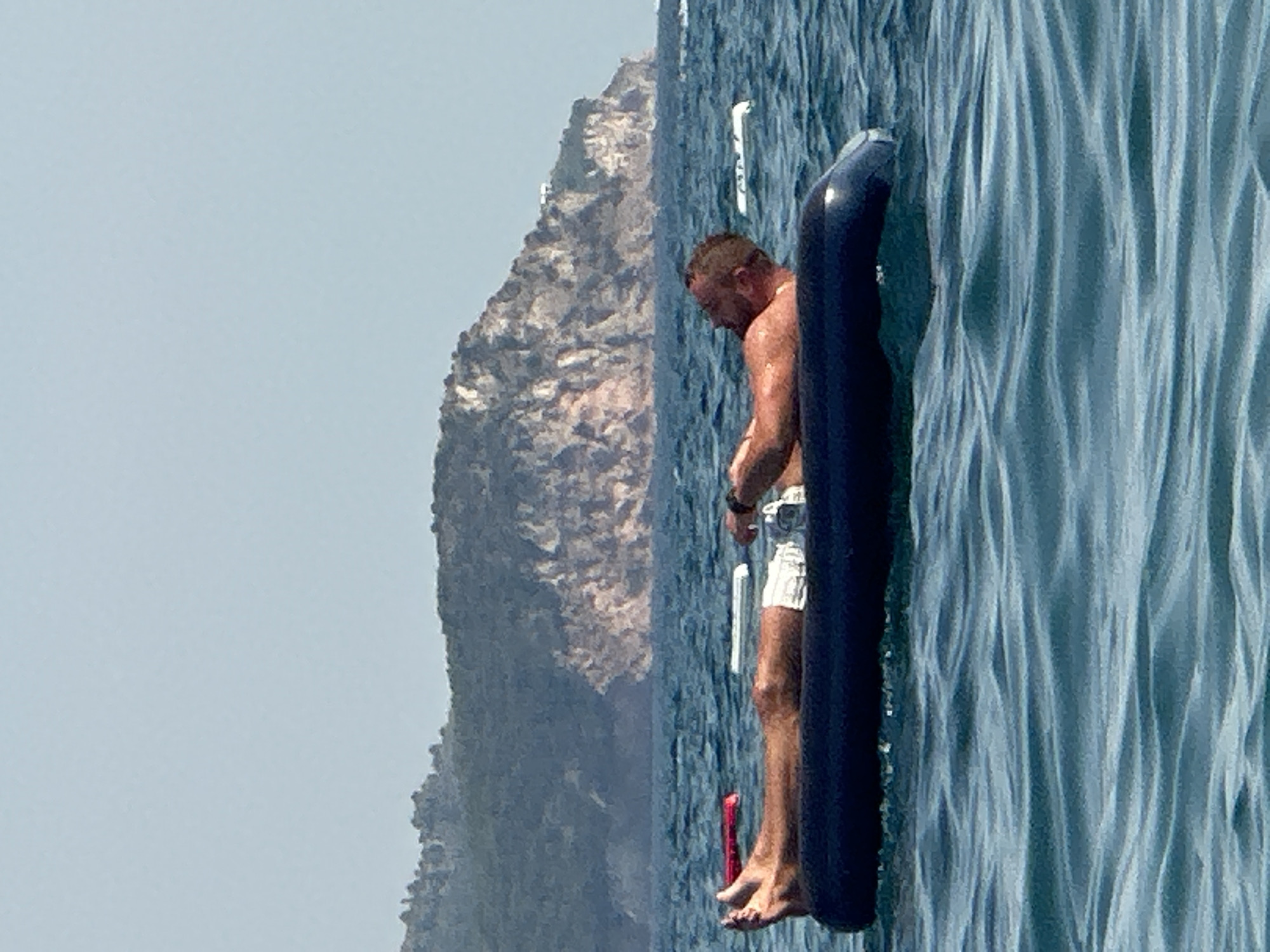 A person is relaxing on an inflatable raft in the sea with a mountainous island in the background. The person is lying on their back, with their arms resting on their chest. They are wearing white swim shorts. The raft is dark blue. The sea is calm, with gentle waves. The sky is clear and blue. The island in the background is covered in green vegetation and has rocky cliffs. The scene appears to be a sunny day in a coastal area, possibly near Boreti, Montenegro, given the typical landscape found there.
