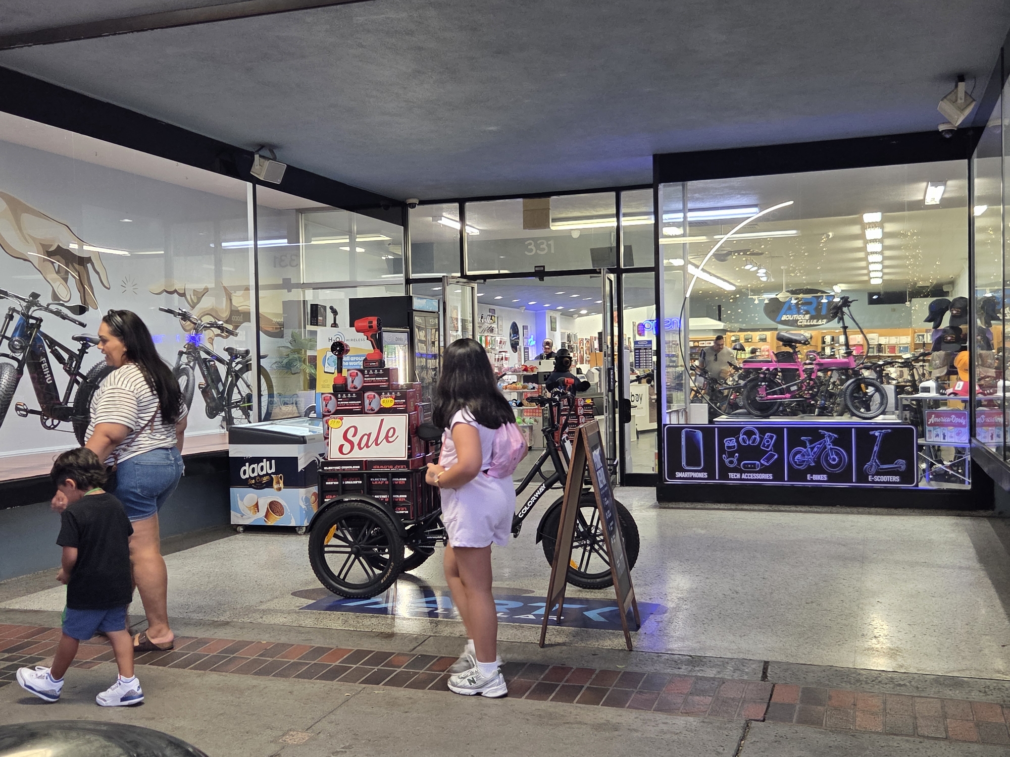 The image captures a street-level view of a commercial storefront in Burbank, United States of America. The storefront features large glass windows displaying various items for sale. In the foreground, a young boy in a black t-shirt and blue shorts walks on a paved walkway with a brick border. To his right, a woman in a striped t-shirt and denim shorts stands near a three-wheeled electric bicycle with a "SALE" sign. Behind her, an ice cream freezer is visible. Further into the image, inside the store, several electric bicycles and scooters are displayed. A woman in a light pink outfit is standing in front of the electric bicycle, looking towards the store. The store's interior is brightly lit, with shelves of products visible in the background. A sign above the entrance reads "331". The storefront also displays icons representing "SMARTPHONES", "TECH ACCESSORIES", "E-BIKES", and "E-SCOOTERS". Security cameras are mounted on the exterior of the building.