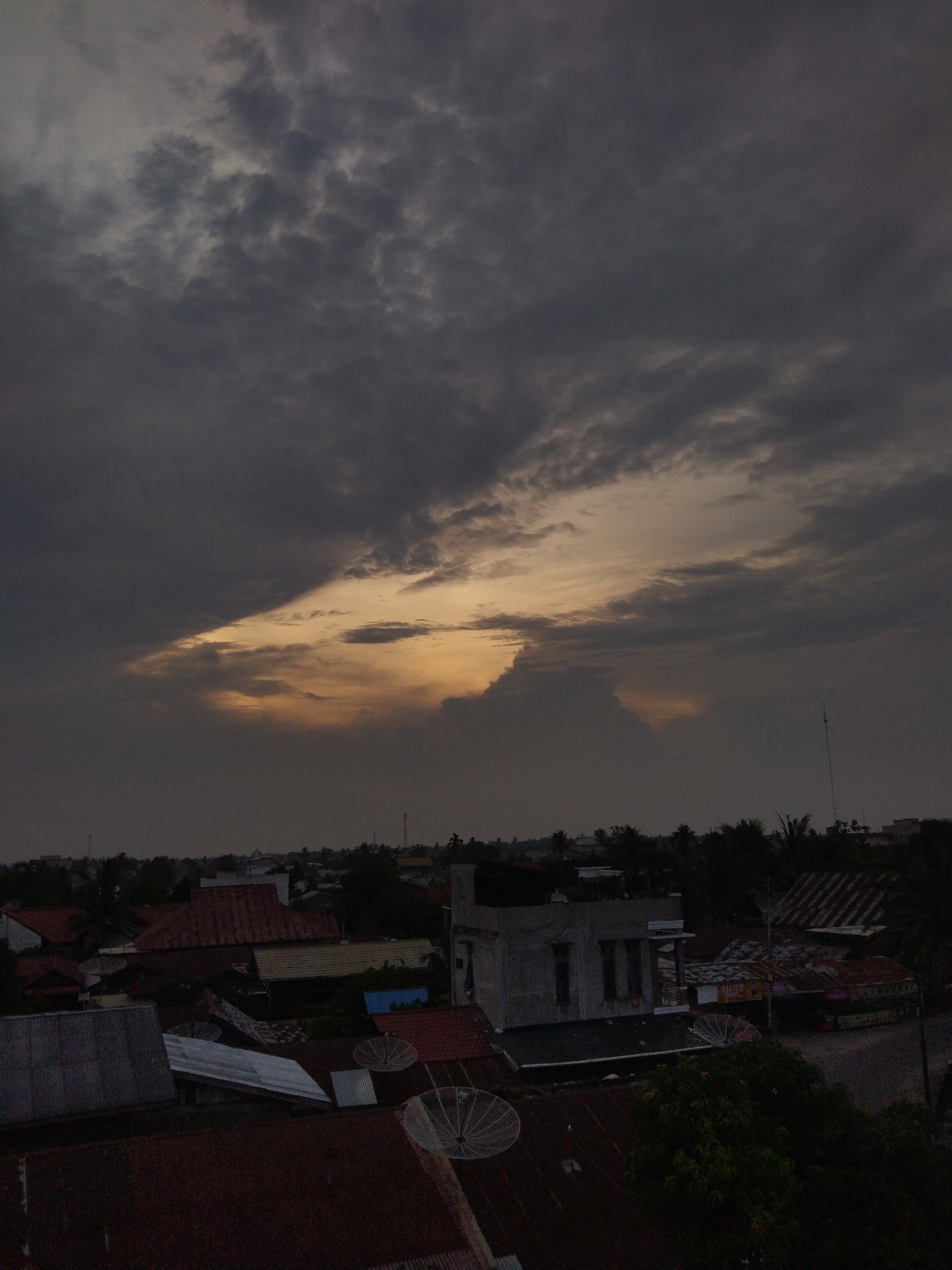 The media file presents an aerial view of an urban or semi-urban landscape under a dramatic, overcast sky. The sky is largely covered by heavy, dark clouds, but a prominent break in the cloud cover reveals a striking golden-orange light, indicative of either sunrise or sunset.
Below the horizon, the scene is dominated by a cluster of buildings, primarily showing their rooftops. These roofs vary in material, including reddish tiles and corrugated metal sheets. Several white, circular satellite dishes are distinctly visible on multiple rooftops, suggesting their common use in the area. Scattered among the buildings and extending into the background are numerous green trees, some appearing to be palm trees. On the far right, a tall, thin antenna or mast rises above the cityscape.
No people are visible in the image, and no specific activity is taking place other than the natural progression of twilight. The time of day is clearly either dawn or dusk, characterized by the low, warm light piercing through the dark clouds. The overall impression is moody and serene, with the golden light providing a focal point. The architectural style, dense housing, and tropical vegetation are consistent with the provided location context of Indonesia. While some faint text might be present on a distant sign, it is too blurry to be legibly identified.