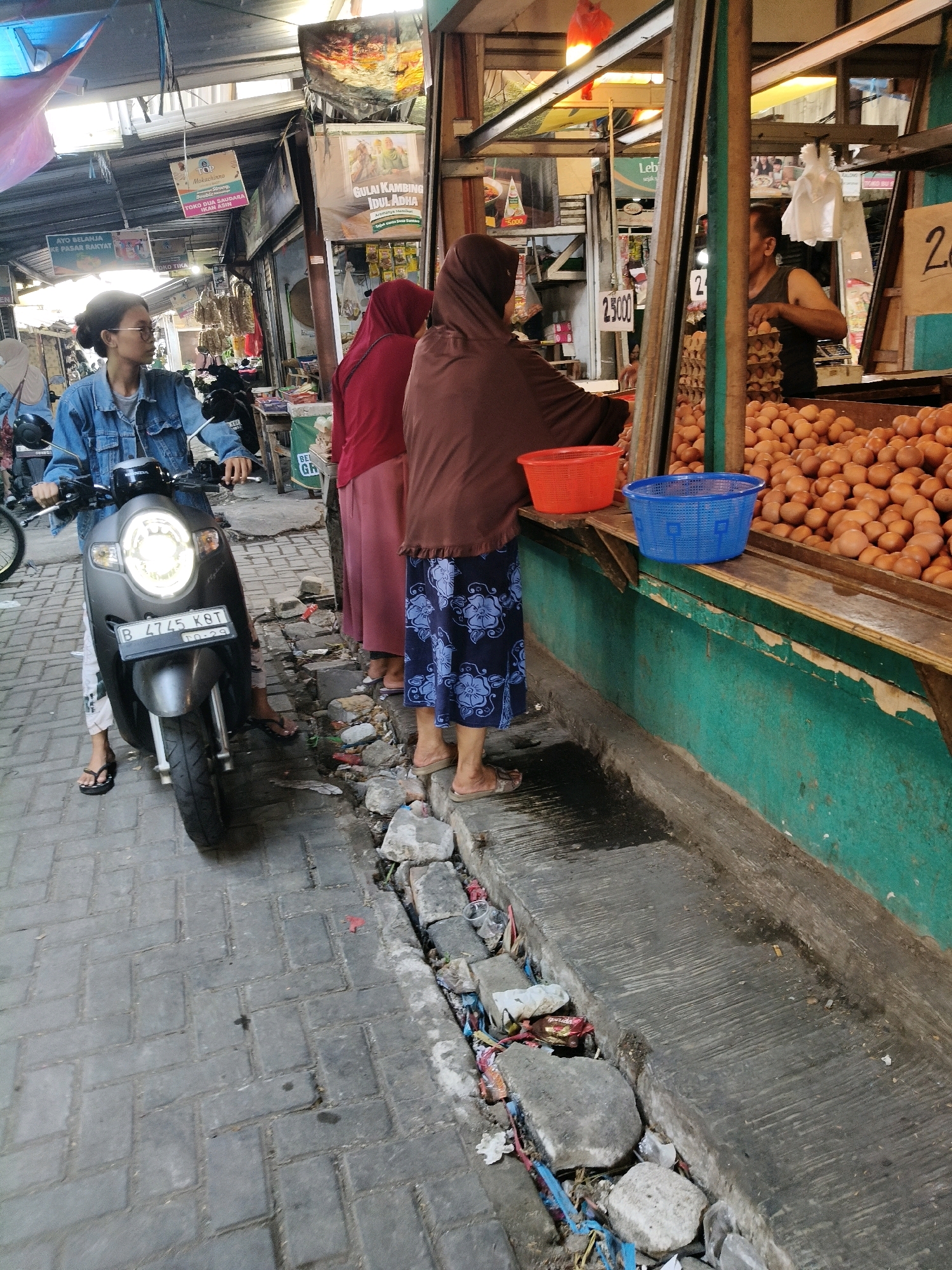 A street market scene in Setiamekar, Indonesia, features a young woman on a scooter, three women standing at a vendor stall, and a large pile of eggs. The woman on the scooter is wearing a denim jacket and glasses, and her scooter's headlight is illuminated. She is positioned on the left side of the frame, facing towards the right. To her right, three women are standing near a market stall. Two of them are wearing hijabs, one in red and one in brown. The woman in brown is wearing a blue floral skirt, and the woman in red is wearing a pink skirt. They are interacting with the vendor, who is partially visible behind a large display of eggs. The stall is painted green and has a wooden counter displaying two baskets, one red and one blue, and a large quantity of brown eggs. The ground is paved with interlocking gray bricks, and a drainage ditch filled with debris and some standing water runs along the side of the stall. Various market signs are visible in the background.