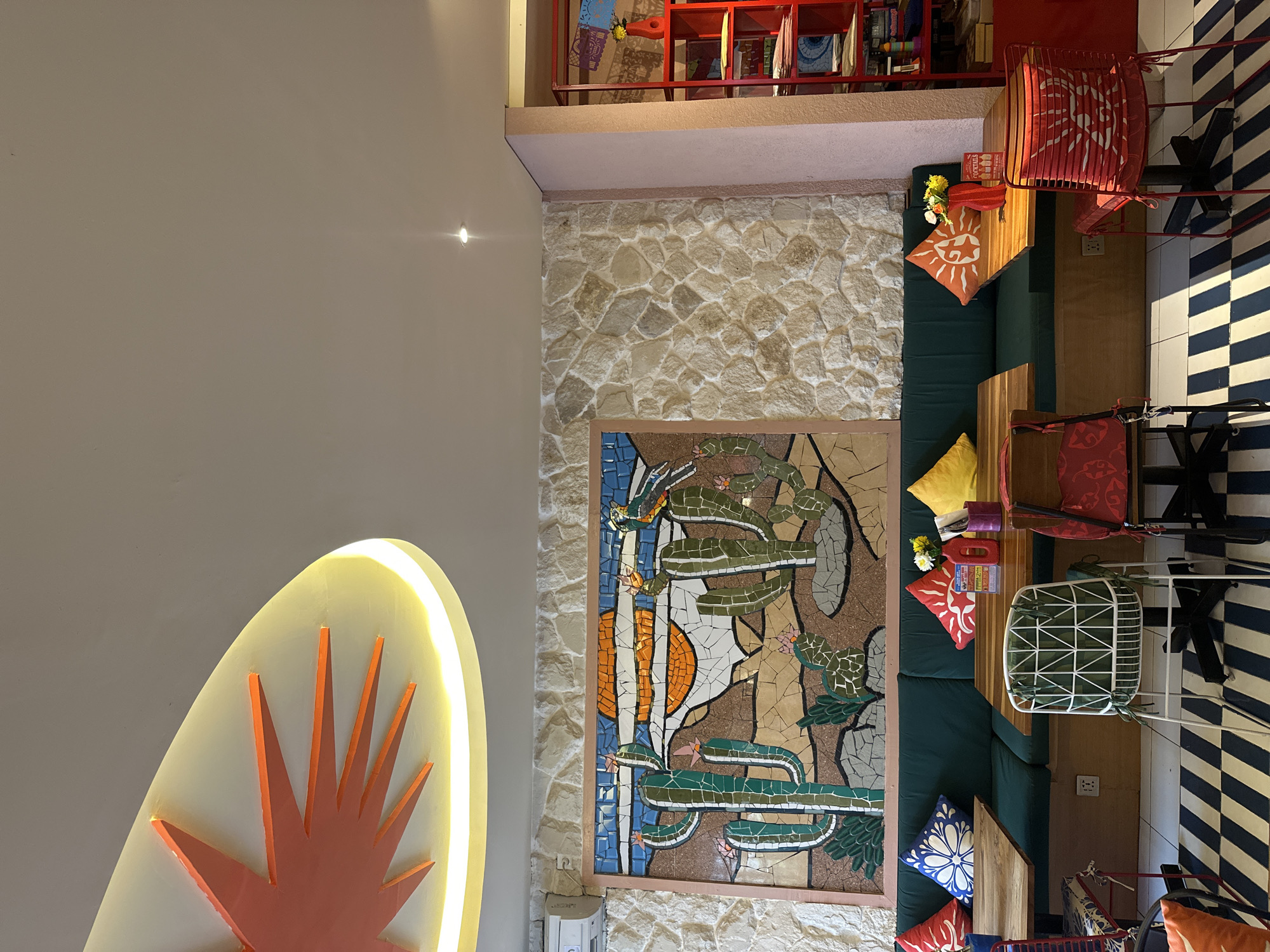 The image depicts the interior of a restaurant in North Kuta, Indonesia, likely a Mexican-themed establishment given the mosaic artwork. The scene is set with a long, dark green banquette seating area against a stone-tiled wall. Above the banquette, a colorful mosaic mural features cacti, a desert landscape, and birds under a sunset. Wooden tables are placed in front of the banquette, some set with decorative pillows in vibrant colors like red, blue, and yellow, along with a red vase holding a small bouquet of flowers.
Several chairs are visible around the tables. One prominent white metal chair with a geometric pattern on its seat is in the foreground, with other chairs, including red metal ones with patterned seats, flanking it. The floor is covered with a striking black and white striped rug. To the right of the banquette, a bright red shelving unit holds various items, including books and colorful decorations. The ceiling features an unusual, abstract light fixture with orange rays radiating from a central point. The overall atmosphere is lively and colorful, suggesting a casual dining environment. No people are present in the image.