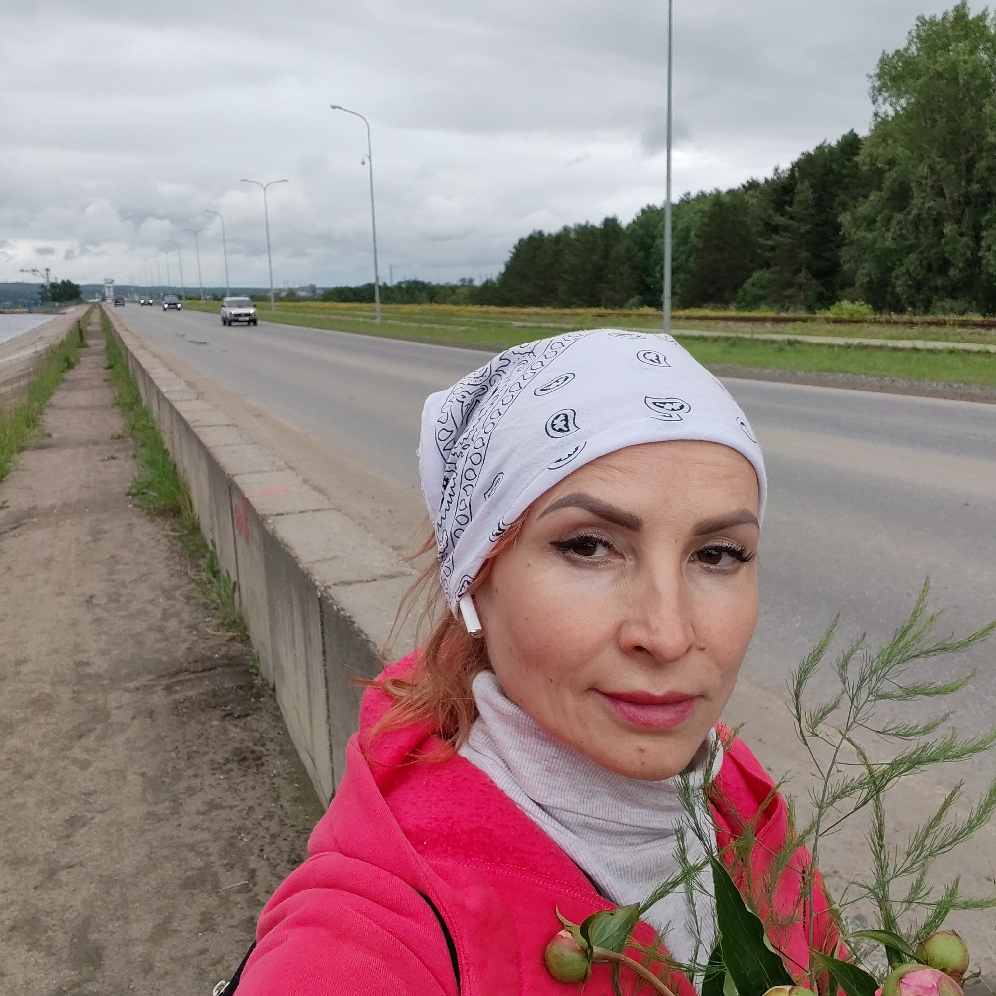 An adult female is depicted in a selfie taken outdoors on an overcast day in Chaikovsky, Russia. She has brown eyes, eyeliner, and long eyelashes, wearing a white bandana with black paisley patterns, a bright pink hooded jacket, and a light grey ribbed turtleneck. A white wireless earbud is present in her left ear. She holds a bunch of green, fern-like foliage with several closed pink peony buds. The background features an asphalt road with multiple lanes extending into the distance, carrying several vehicles, including a white SUV. A series of tall, double-armed streetlights line the road. To the immediate left of the subject is a concrete pedestrian path running alongside a concrete barrier. Beyond this barrier, a body of water is partially visible, with a large crane structure in the far distance. To the right of the road, a grassy verge transitions into a dense forest of coniferous trees, with railway tracks discernible between the road and the treeline. Further beyond the trees, a field of yellow-flowering plants is visible under a uniformly cloudy sky.