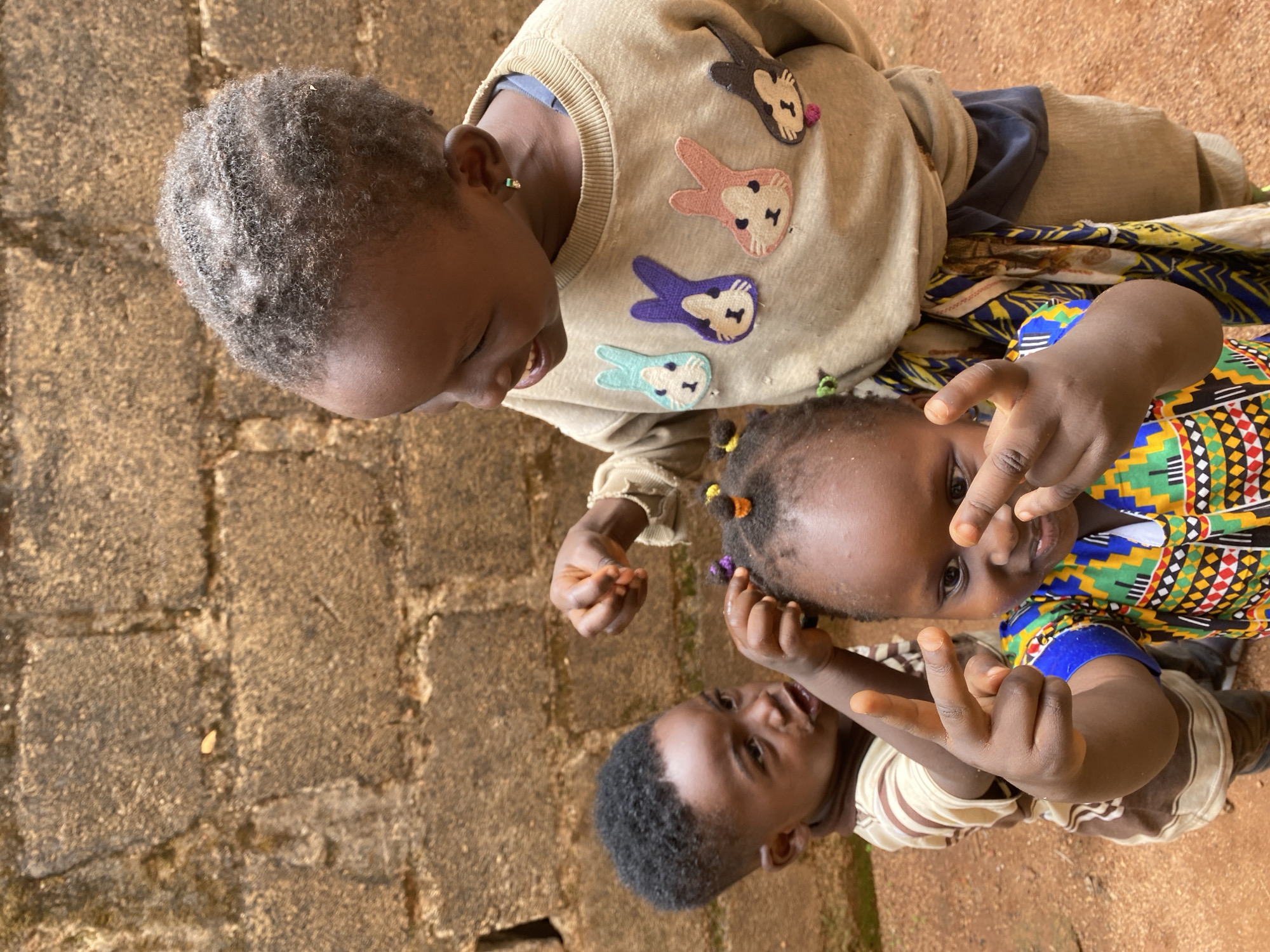 Three young children are captured from an overhead perspective, gathered closely together, possibly sitting or kneeling outdoors on a reddish-brown, earthy ground.
The child in the upper left, appearing slightly older, has short, curly dark hair and wears a light brown long-sleeved top embellished with colorful, embroidered animal appliques—bunnies or cats—in shades of pink, purple, blue, and black. A small green stud earring is visible in their ear. This child looks downwards towards the others with a gentle smile.
To the right, a younger child is prominent, wearing a vibrant shirt with geometric patterns in green, yellow, red, and black, characteristic of West African textiles. This child looks directly at the camera, holding up both hands with two fingers extended in a peace sign gesture. Small colorful ties or beads are noticeable in their dark hair.
Partially visible in the lower left is the youngest child, wearing a light and dark brown striped top. This child is also looking upwards, and one hand is raised, seemingly mirroring the peace sign gesture of the child next to them.
The immediate background is a textured wall made of stones or bricks with visible mortar lines. The scene is illuminated by natural light, indicating daytime. The children appear happy and engaged, suggesting a moment of playful interaction or posing for the camera in Kuru Dungus, Nigeria. There is no visible text in the image.