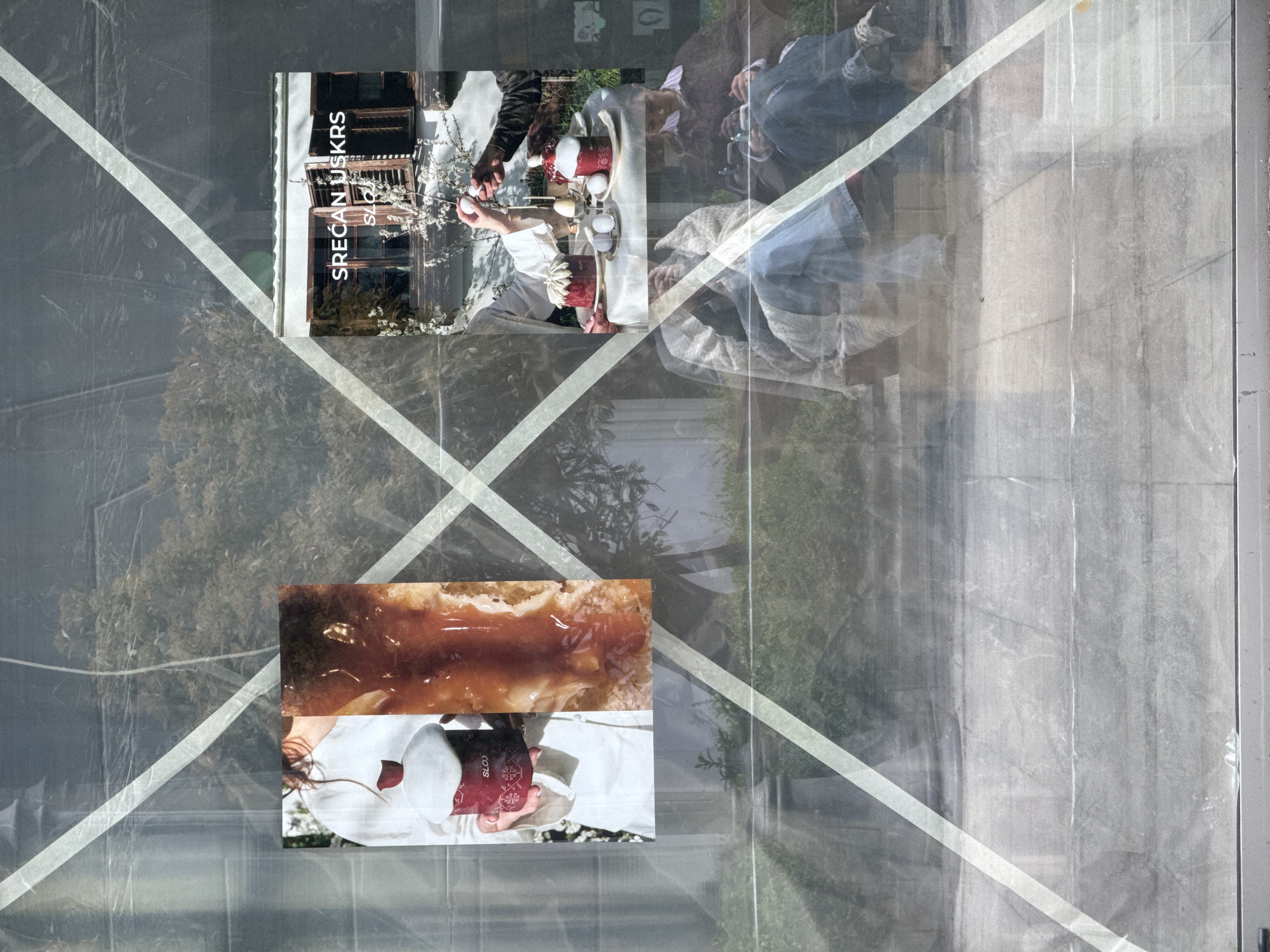 The image appears to be an abstract, artistic photo taken in Belgrade, Serbia. It is difficult to ascertain precise details as the picture contains multiple layers and visual effects. In the foreground, there is a translucent material, possibly plastic, with white diagonal tape, creating an overlaid "X" pattern. Behind this, there are two images of food and people seemingly superimposed over an outdoor scene, where reflections of buildings and greenery can be observed. In the top left, the text "SRECAN USKRS" is visible, which translates to "Happy Easter." The first of the images shows an Easter celebration, with people holding eggs and cake, which are cultural traditions in Serbia. A second image shows a brown sauce atop what seems to be a pastry. The background reflects an urban landscape with trees, parts of structures that appear to be windows and walls, possibly within Belgrade.