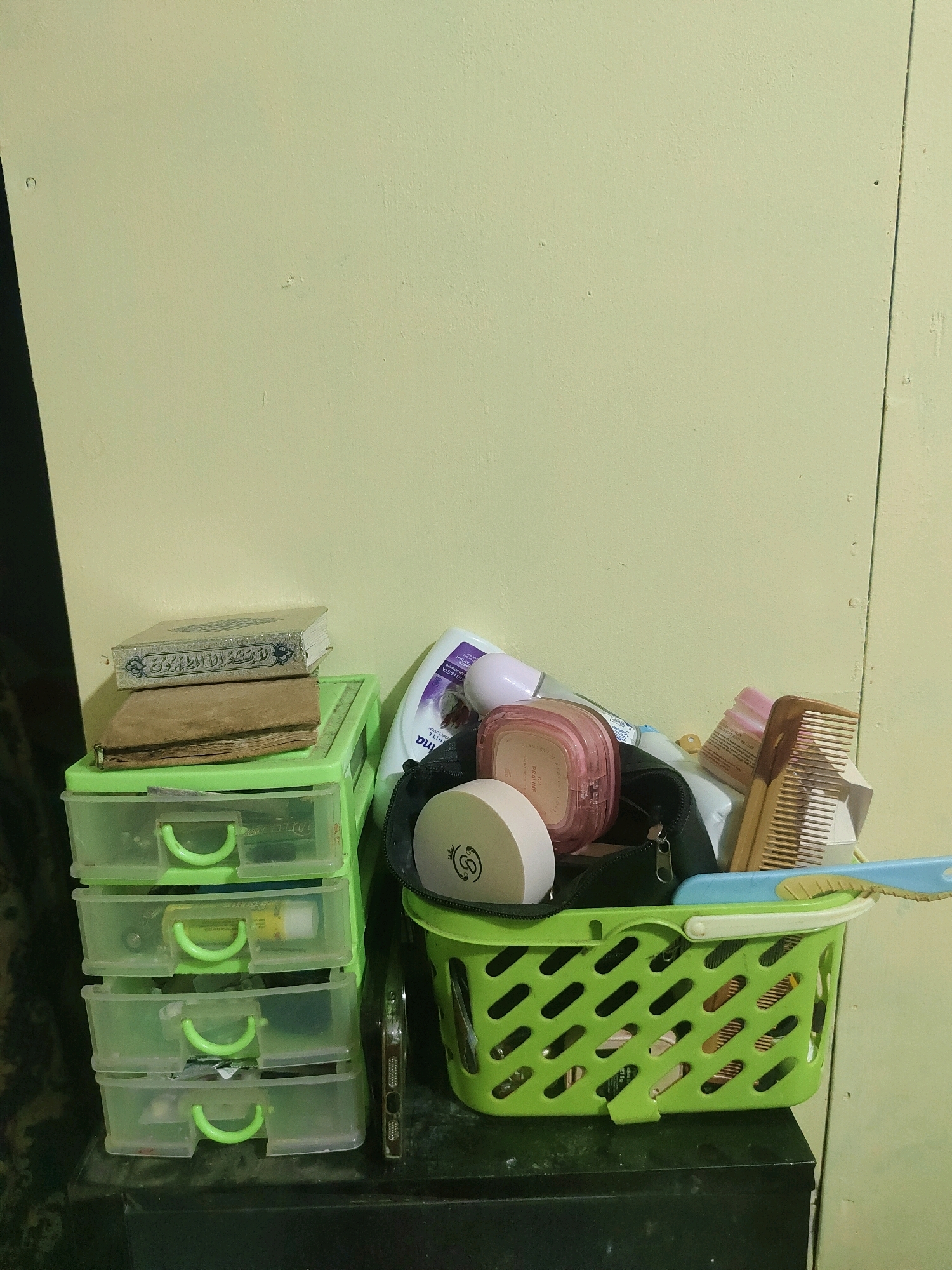 A stack of four transparent green plastic drawers sits on a dark surface, topped by two books, the upper one bound in ornate gold and white fabric, the lower one in brown leather. To the right of the drawers is a green plastic basket filled with various personal care items. These include a pink compact, a bottle of lotion, a wooden comb, and a blue hairbrush. A black makeup bag is also partially visible within the basket. The scene is set against a pale green wall, with a vertical seam or door jamb on the right side. The lighting suggests an indoor environment, likely a room in a home or similar establishment in Sukanagara, Indonesia.