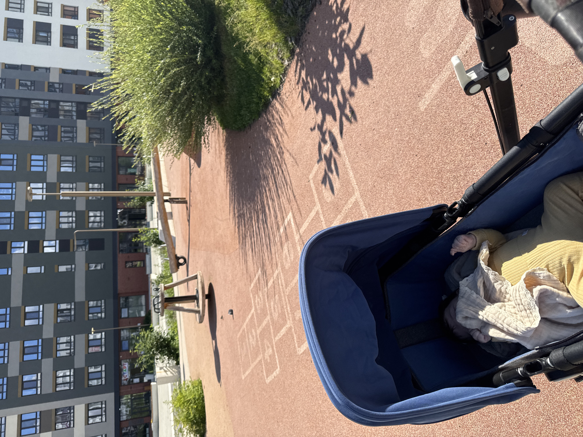 This media file captures a view from a stroller, looking down at a baby and then outwards to a modern apartment building and a playground. The baby, swaddled in a light-colored blanket and wearing a yellow outfit, is the primary subject within the stroller. The stroller itself is dark blue.
The scene is set outdoors on a sunny day, indicated by the distinct shadows of trees cast on the reddish-brown pavement. The playground area features chalk drawings of hopscotch squares. In the background, a multi-story apartment building with a contemporary architectural design dominates the view. The building has a facade with alternating colors of gray, white, and dark panels, featuring numerous windows. Greenery, in the form of bushes and trees, is visible around the building and on the playground.
The lighting suggests it is daytime, likely morning or afternoon, given the strong shadows. The overall mood appears peaceful and domestic, with the focus on the child and the surrounding urban environment. There is a sign visible on the apartment building with Cyrillic text that appears to read "СТАРТ," which translates to "START." The location context provided is Patrusheva, Russia.