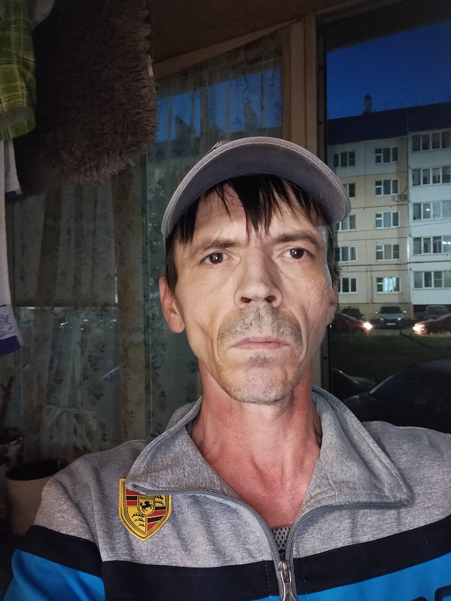 This is a selfie taken by a man outdoors at dusk in Zyuzino, Russia. The man is wearing a grey baseball cap and a grey and blue zippered jacket with a Porsche crest on the left chest. He has a thin build and fair skin, with visible lines on his face and a reddish hue on his neck. His expression is neutral.
In the background, we see a multi-story apartment building with many windows, typical of Russian urban landscapes. Several cars are parked in front of the building, and the sky is a deep blue, indicating it is evening or twilight. The setting appears to be a residential area. There is no explicit indication of any activity or event, other than the man taking a photograph of himself.