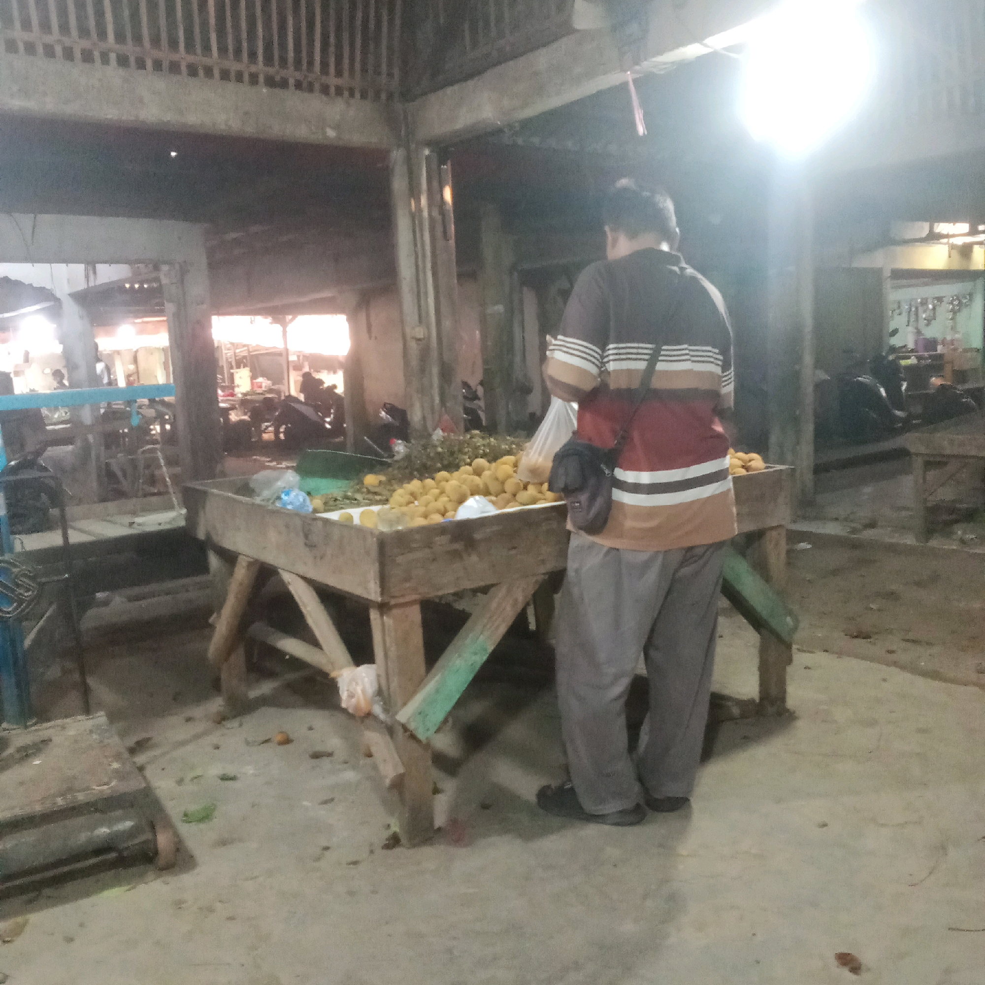 The image depicts an outdoor marketplace scene in Karawang, Indonesia, likely at night or dusk given the artificial lighting. A person, viewed from behind, stands beside a wooden stall laden with a large quantity of round, yellow fruits, possibly oranges or tangerines. The stall is supported by weathered wooden beams, some painted green. The ground is a concrete surface, with scattered debris and some fallen leaves or produce. In the background, there are other stalls, a motorcycle, and the suggestion of a covered structure with vertical supports. The lighting is bright and uneven, with a strong source creating a glare in the upper right portion of the image. The person is wearing a striped shirt with maroon, brown, and white bands, and grey trousers, with a dark bag slung across their body. The overall impression is of a busy, yet somewhat dimly lit, local market environment.