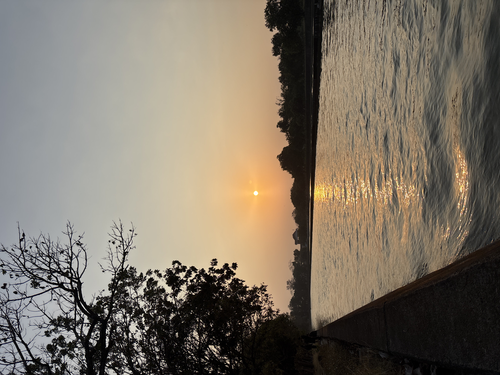 The image shows a sunset over a body of water in Beber, Indonesia. A tree with bare branches is silhouetted on the left side of the frame, with some leaves still attached to the lower branches. The sun, a bright orange orb, is setting in the distance, casting a golden reflection on the rippling water. The opposite bank is lined with trees and some buildings are visible in the background. A concrete embankment runs along the left edge of the water, with a few people walking along it. The sky is a gradient of pale blue and orange, typical of a sunset.