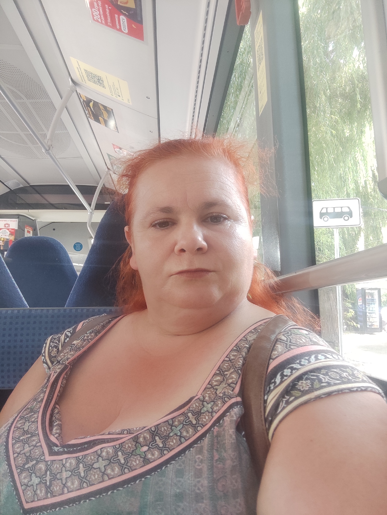 A woman with reddish hair is taking a selfie on a bus in Chernivtsi, Ukraine. She is wearing a patterned dress and has a brown strap across her shoulder. The interior of the bus is visible, including blue seats and a sign on the ceiling. Through the window, trees and a bus stop sign can be seen. A Pepsi refrigerator is also visible outside the bus.