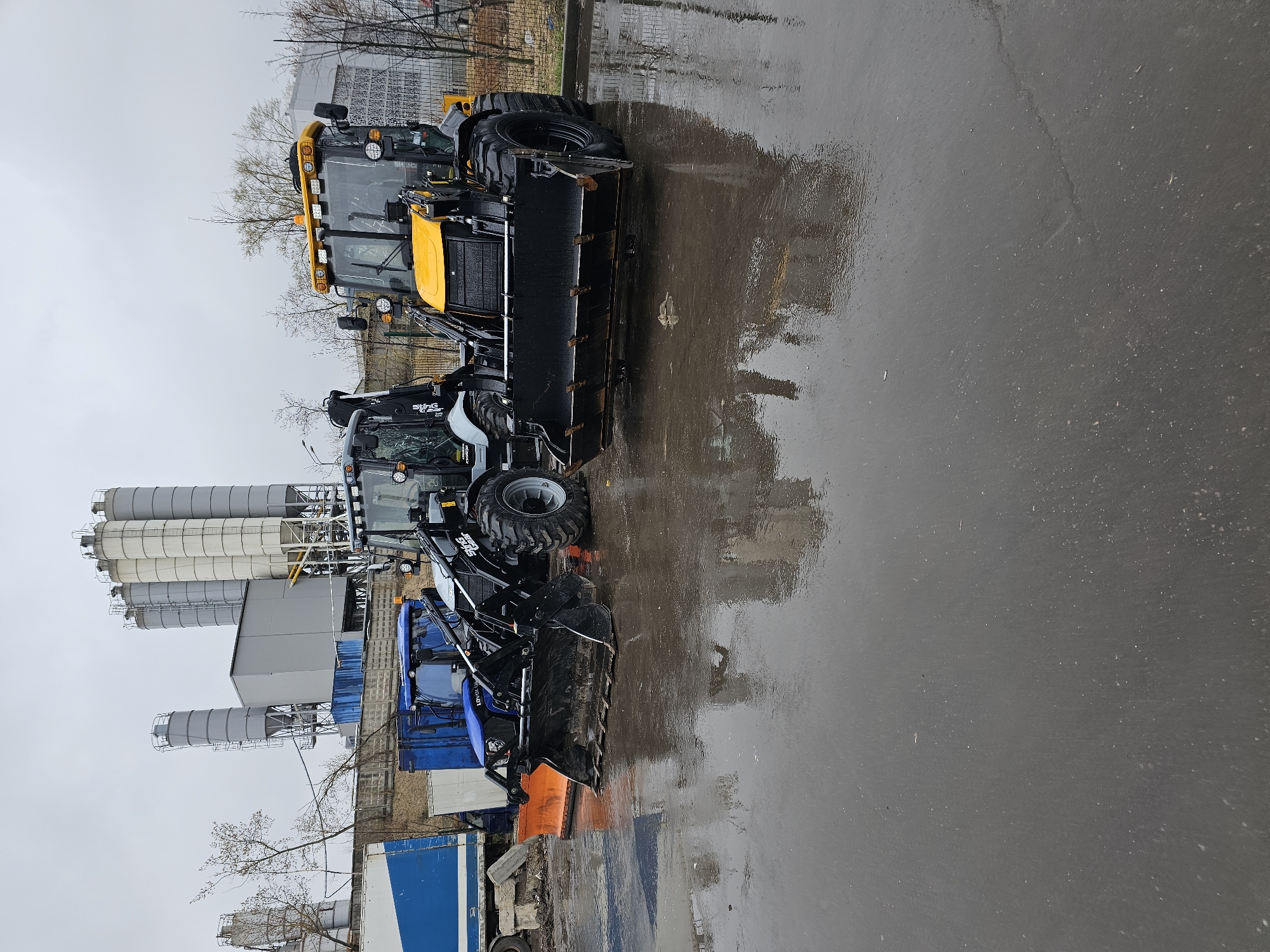 The scene depicts a wet, likely industrial or construction yard. Three backhoe loaders are parked in a row on a gray, reflective surface, suggesting it is recently rained.
The first backhoe, furthest from the viewer, is primarily blue with an orange snowplow attachment. The middle backhoe is painted in shades of gray and black, while the backhoe closest to the viewer is predominantly yellow.
In the background, there are several large, cylindrical silos, typical of concrete or cement plants. There are also some trailers, and a fence.
The location is Khimki, Russia. This is indicated by the presence of what appears to be industrial equipment commonly found in manufacturing or material processing facilities, and the weather and architecture are consistent with that area.