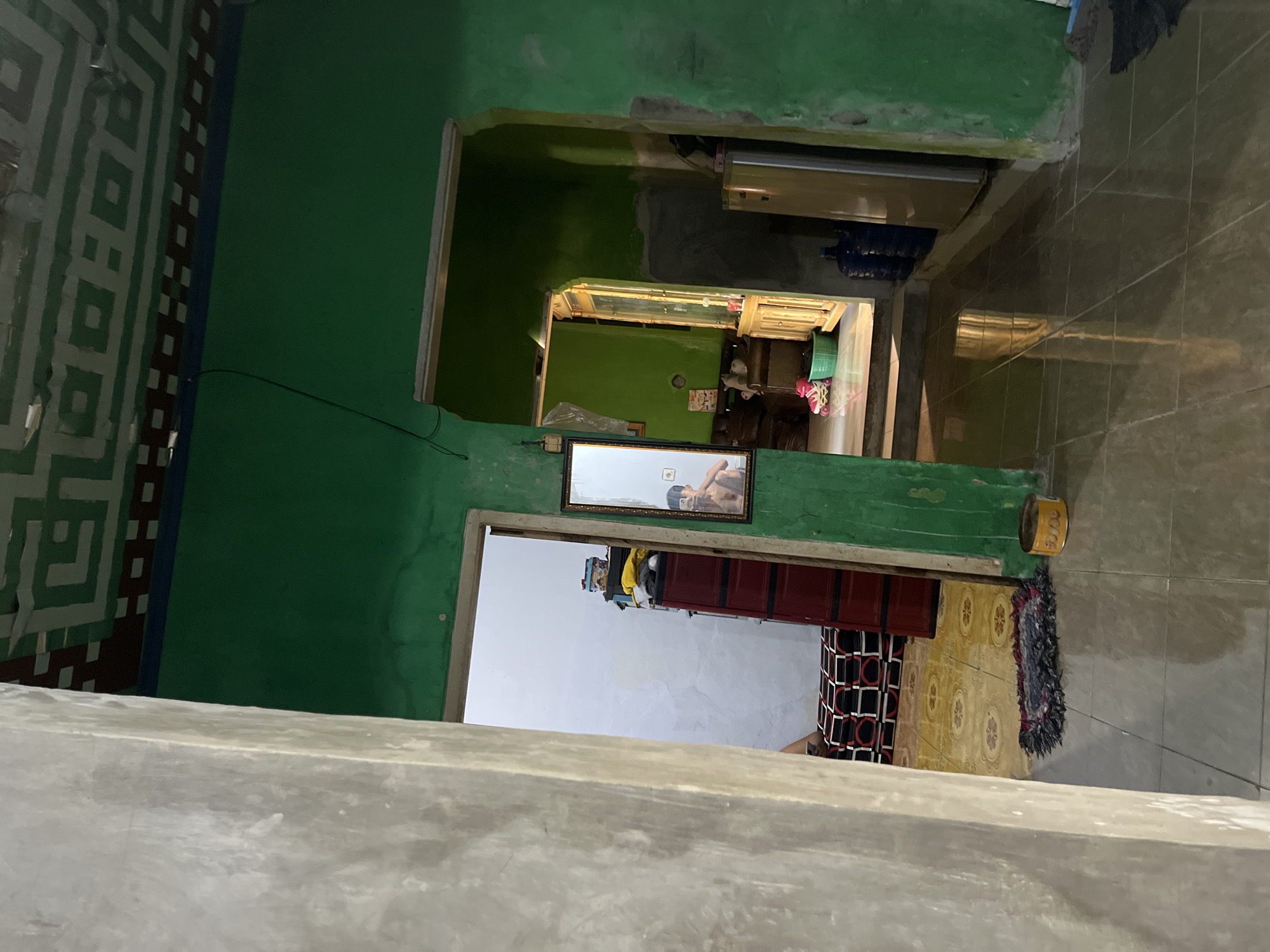 The image captures an interior scene within a residence in Laban, Indonesia. A person is visible in a mirror reflecting their image. The room features green walls and a patterned ceiling. Objects in the scene include a red plastic cabinet, a refrigerator, water jugs, a small rug, and roller skates. The floor is tiled and reflects light. The overall impression is of a lived-in, somewhat cluttered domestic space.