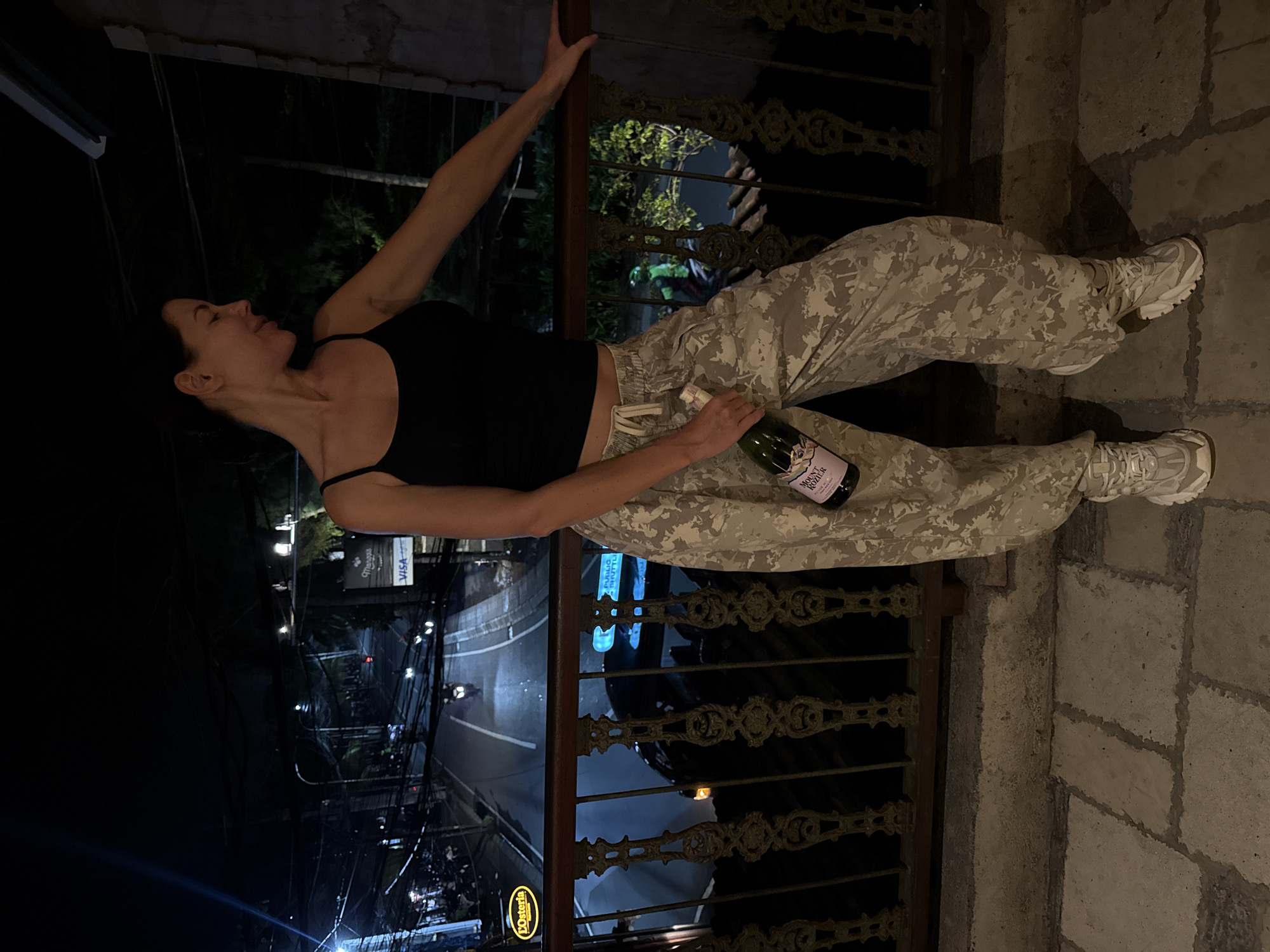 A woman with dark hair, wearing a black tank top and beige camouflage pants, is lying on a wrought-iron balcony at night. She holds a bottle of wine in her right hand. The balcony overlooks a street with traffic, identified by signs for "VISA" and "LOsteria," in Unknown, Unknown. The background features dark buildings and overhead power lines. The balcony's floor is made of stone.