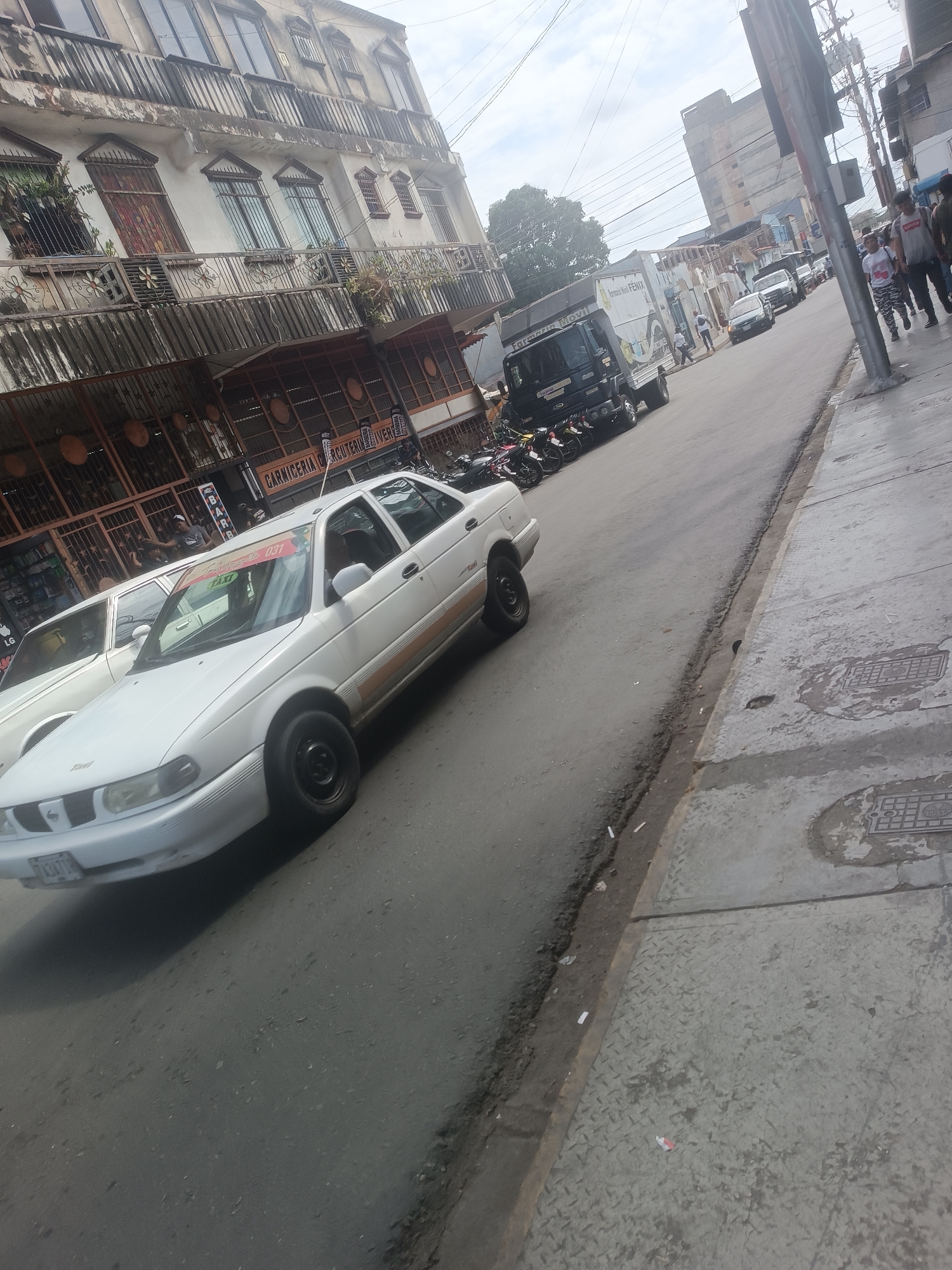 This image captures a street scene in Porlamar, Venezuela, depicting a view looking down a street with buildings lining the left side and a sidewalk on the right.
The street is an asphalt two-lane road with a visible curb separating it from a concrete sidewalk on the right. The sidewalk contains two rectangular metal manhole covers and shows signs of wear and debris. The leftmost side of the image features a multi-story building with a deteriorated facade, multiple windows, and balconies on its upper floors. The ground floor of this building houses commercial establishments, including one displaying a sign partially visible as "CARNICERIA CHACUTERIA Y VIVERES".
Several vehicles are present. In the foreground, a white four-door sedan (Nissan Sentra B13 model, common as a taxi in Venezuela) is parked or moving slowly. It has a visible license plate "A 13AVJ" and a red banner on its windshield with "TAXI 037" printed on it. Its rear passenger door has a horizontal strip of light brown trim. Another white sedan is partially visible behind it. Further down the street, a black light-duty truck with a white box on its bed displays "Farmacia Movil FENIX" on its side. More vehicles, including cars and several motorcycles parked along the curb, are visible further in the background.
Multiple individuals are present. On the far right sidewalk, several pedestrians are walking towards the viewer, including one person in a grey t-shirt and another in a patterned top. Other individuals are seen near the shops on the left side of the street and walking near the truck in the mid-ground. The sky above is overcast.