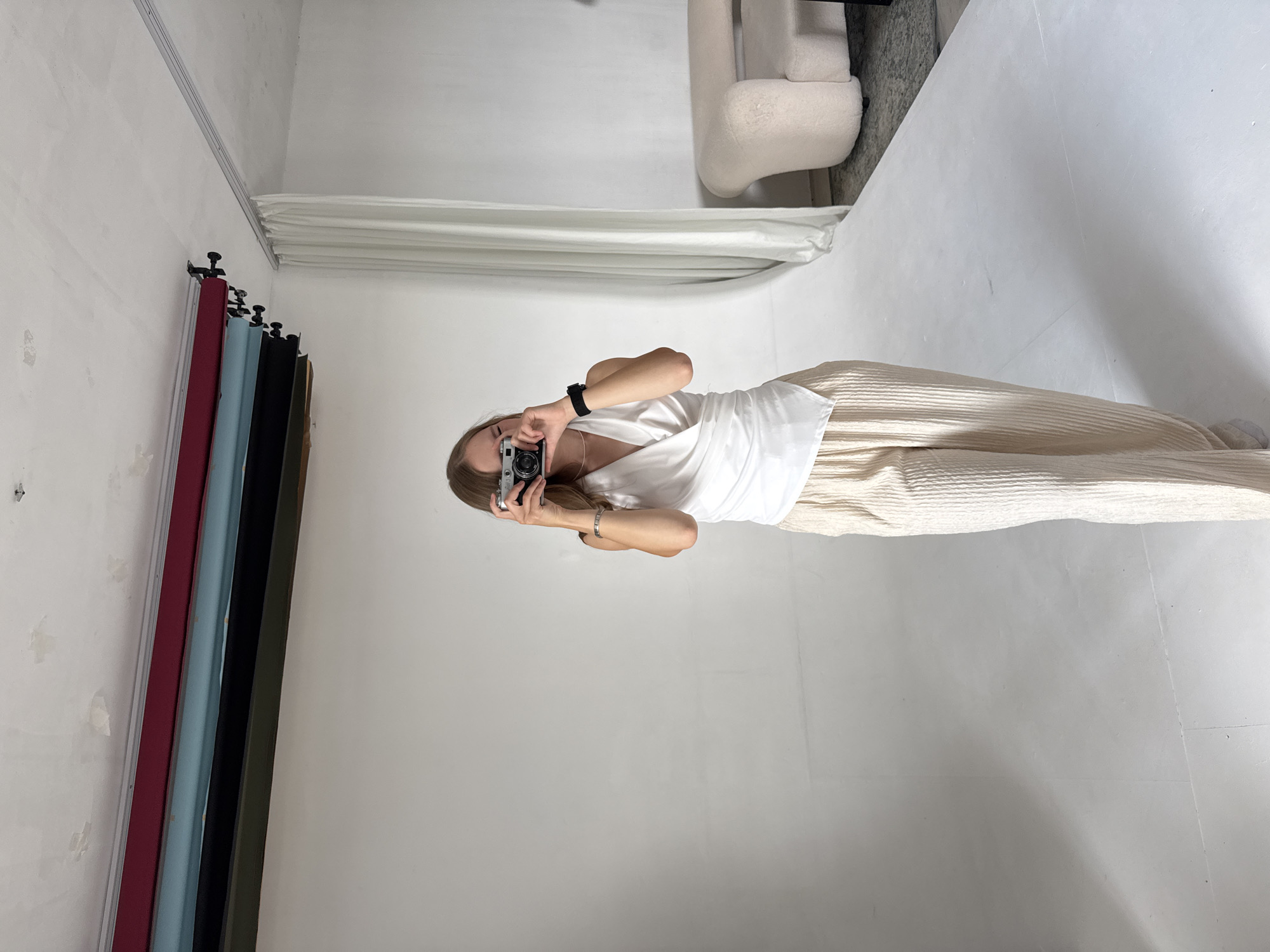 A young woman stands in a studio setting, holding a camera to her eye as if taking a photo. She is dressed in a white wrap top and cream-colored, wide-legged corduroy pants. In the background, there is a white cyclorama, a rack of backdrops (red, blue, and black), and a cream-colored, plush sofa. The ceiling has exposed pipes and some evidence of repairs. The lighting is bright and even, typical of a studio environment. There is no specific indication of the city of Chaikovsky, Russia.