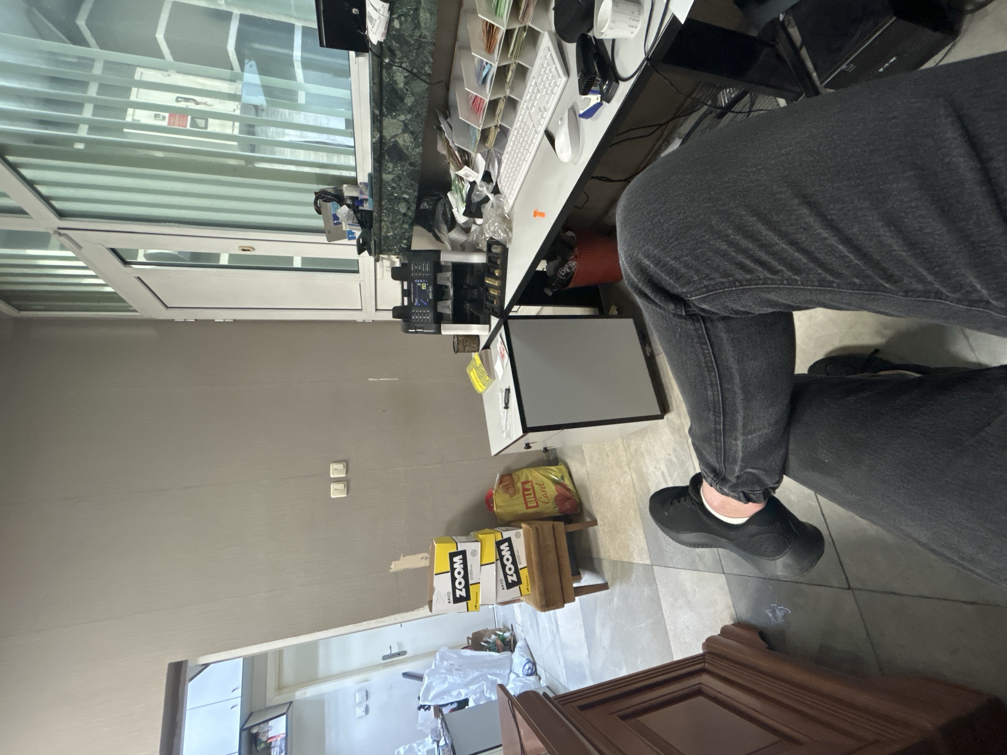 The image appears to be taken inside a business, possibly an office or retail space, in Shumen, Bulgaria. A person wearing dark gray jeans and black sneakers is seated at a desk. The desk is cluttered with office supplies, including a computer keyboard and mouse, papers, and what appears to be a currency counter. Under the desk are boxes of A4 paper, bearing "Zoom" branding, and a bag labeled "Billa Card." A monitor is also present, partially obscured. In the background, there is a window with horizontal blinds, and a glimpse into what seems to be another room with white walls and some furniture.