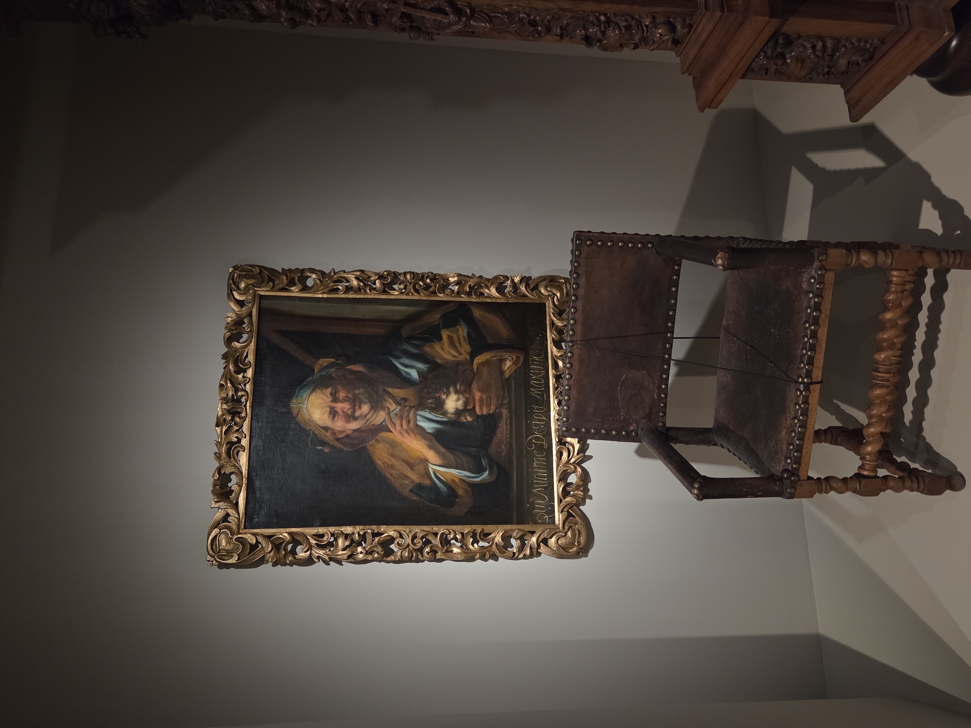 The image shows an interior scene at Malmö Konstmuseum in Malmö, Sweden. In the center of the frame, a painting in an ornate gold frame hangs on a grey wall. The painting depicts an elderly woman wearing a blue headscarf and a yellow and blue garment, holding a small animal in her arms. Below the painting, a dark wooden chair with leather upholstery and decorative metal studs is positioned. To the right of the painting and chair, a portion of a richly carved wooden cabinet or furniture piece is visible. The lighting in the room creates shadows on the wall and floor.