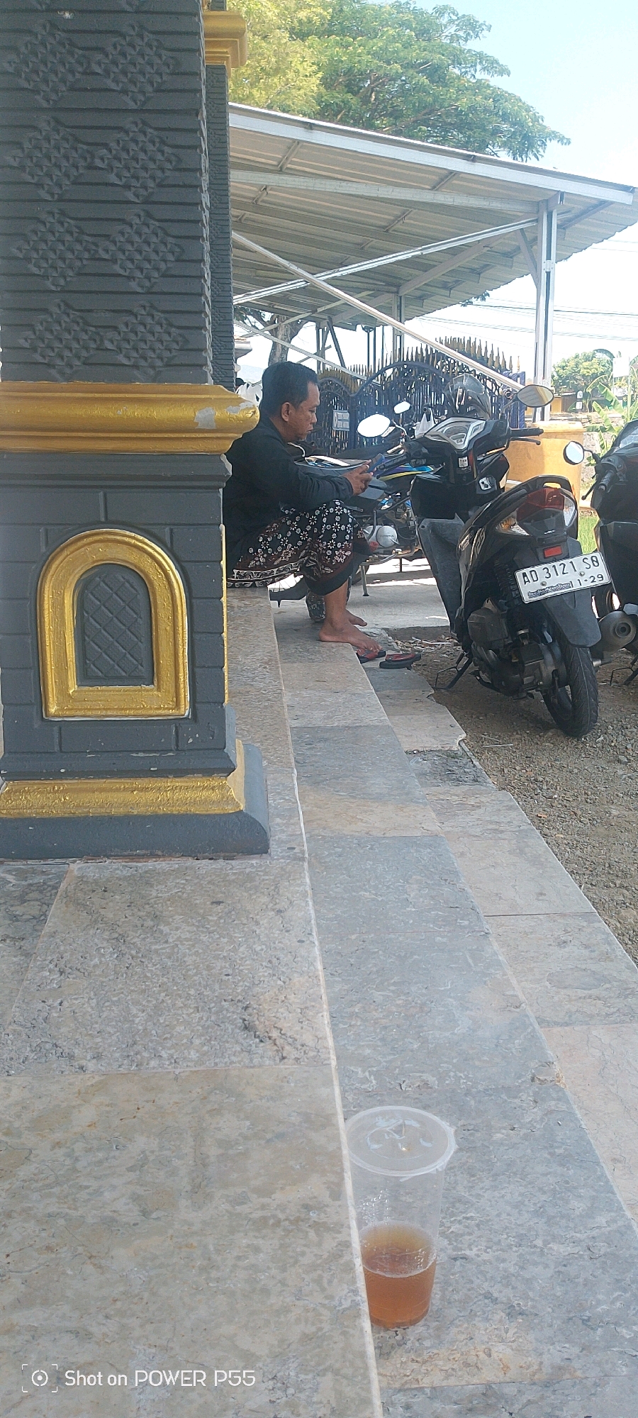 In the city of Weru, Indonesia, a man sits on a concrete step, looking at his phone. He is wearing a black long-sleeved shirt and patterned pants, and is barefoot. To his right, a black motorcycle is parked. The man is positioned on the steps of a building with a dark grey and gold facade. The steps are made of concrete and are slightly worn. A clear plastic cup containing a brown liquid is placed on one of the steps in the foreground. The background reveals a covered outdoor area with a metal roof supported by pillars, and trees with green foliage are visible overhead. The overall scene appears to be an outdoor public space, possibly near a place of worship or a community building.