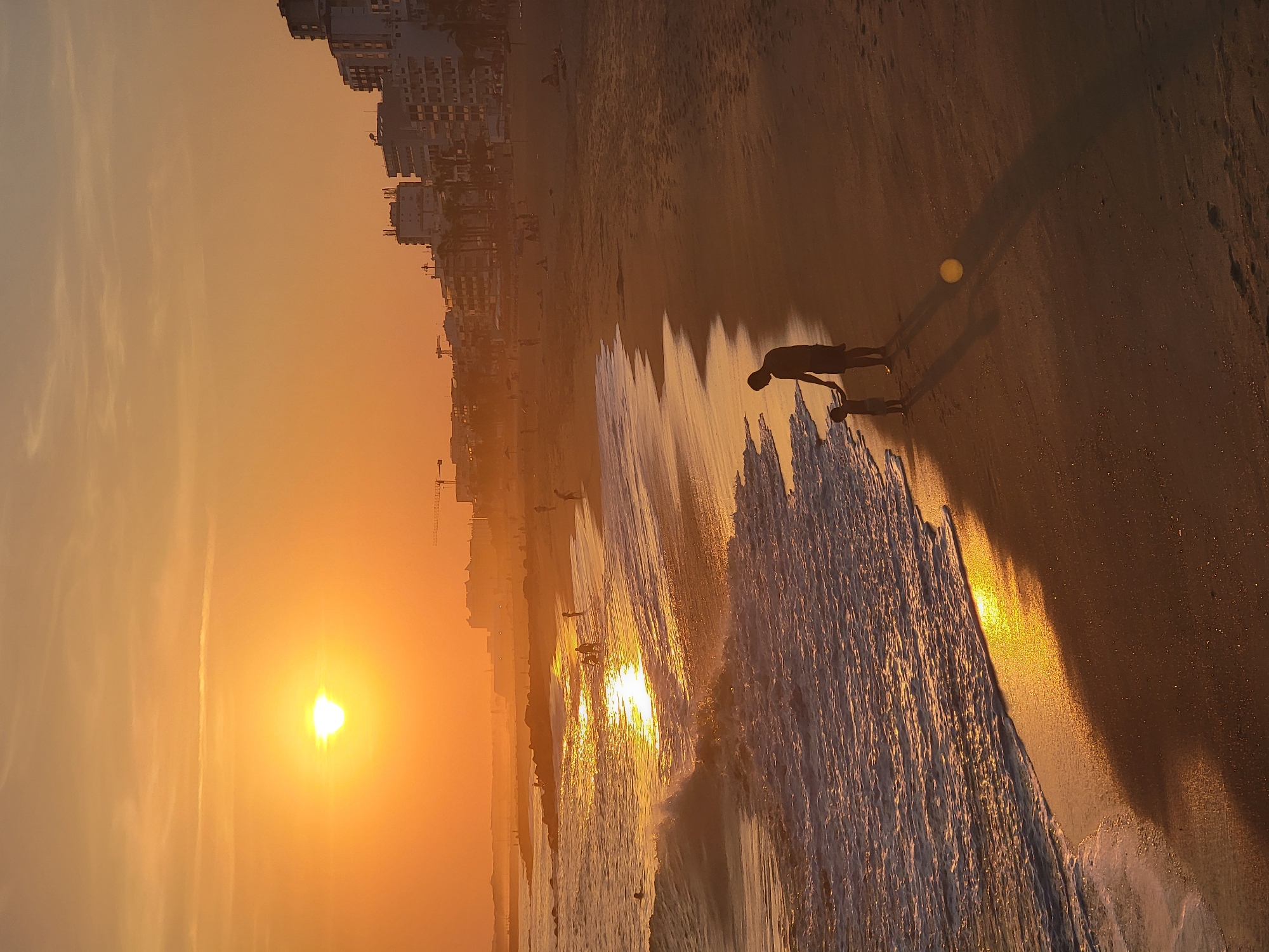 A wide-angle, vertical, high-angle shot captures a sunset on the beach in Quarteira, Portugal. The sun, a bright orb of yellow and white, is low in the sky, casting a warm, orange glow over the scene. The sky is a soft gradient of orange and yellow, with faint wisps of clouds. In the distance, the city skyline of Quarteira is silhouetted against the sky, with buildings of varying heights and construction cranes visible.
The beach itself is wide and sandy, with the gentle waves of the Atlantic Ocean lapping at the shore. The water reflects the golden light of the sunset, creating a shimmering path across the sea. Scattered along the beach are several figures, some wading in the shallow water, others walking along the shore.
In the foreground, a man and a young child stand on the wet sand at the water's edge. The man, silhouetted against the setting sun, is holding the child's hand. Their shadows stretch long across the sand. The child appears to be looking out at the ocean. The overall atmosphere is one of tranquility and natural beauty.