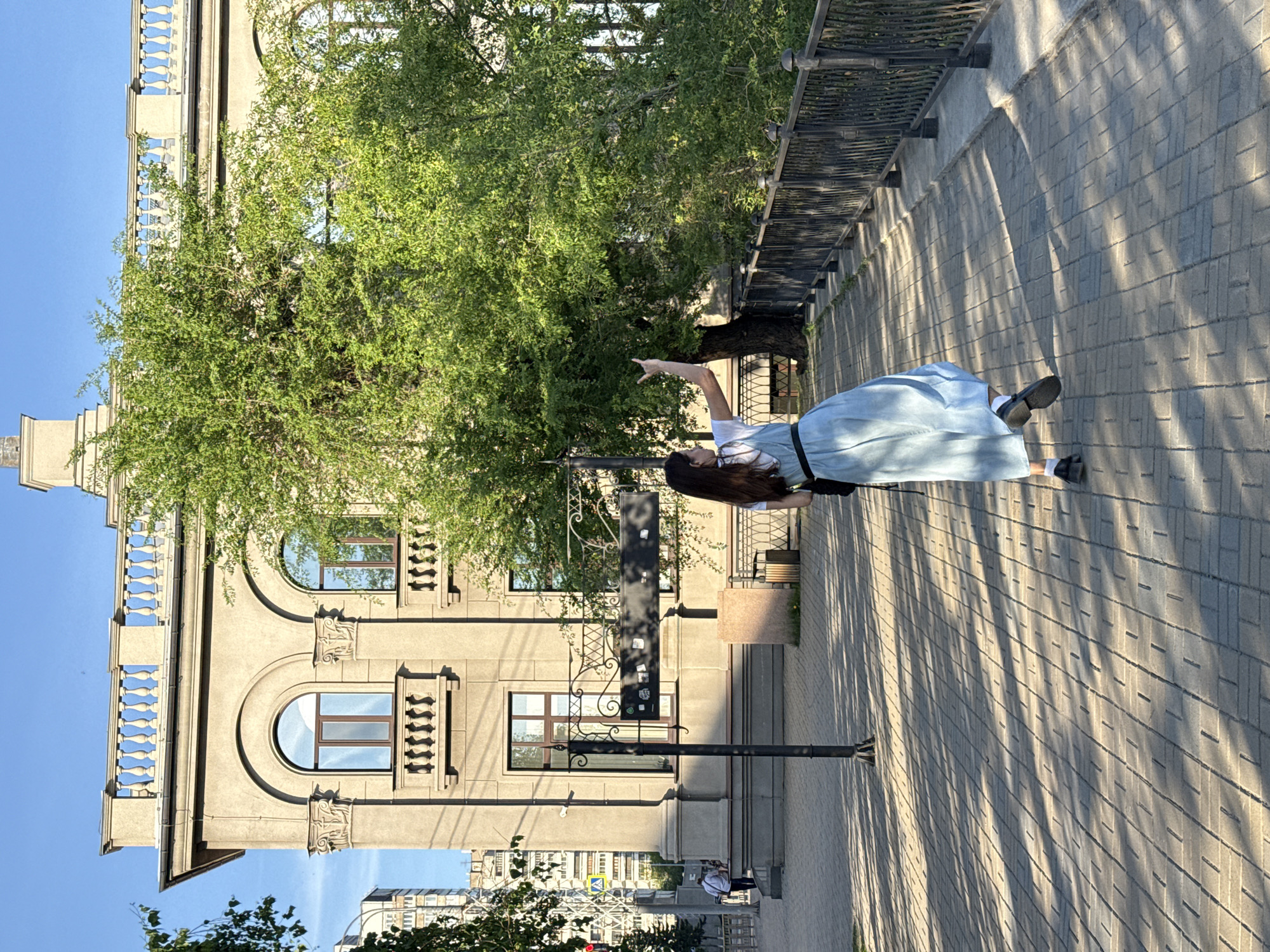 This image depicts a bright, sunny urban scene, likely captured during late morning or early afternoon given the distinct shadows. The central subject is a young woman with long dark hair, seen from behind and slightly to the side. She is dressed in a light blue pinafore or dress over a white top, paired with dark shoes and white socks. Her right arm is raised, with her index finger pointing upwards and to the left, seemingly indicating something to her unseen companion or viewer.
She stands on a neatly paved sidewalk, which features a distinctive rectangular pattern, partially covered by long, diagonal shadows. To her right, a dark metal fence or railing runs parallel to the sidewalk. A large, vibrant green tree with dense foliage occupies a significant portion of the left side of the frame, partially obscuring the building behind it.
The background is dominated by an impressive, multi-story building with a light-colored facade, showcasing classical architectural elements such as decorative cornices, arched windows with dark frames, and balconies featuring ornate balustrades. A black lamppost or street sign pole stands near the woman, bearing a few small, unidentifiable rectangular stickers. In the far distance, other city buildings are visible under a clear blue sky. The scene conveys a pleasant, daytime street atmosphere in an urban environment, specifically Krasnoyarsk, Russia, as indicated by the context. No readable text is visible in the image.