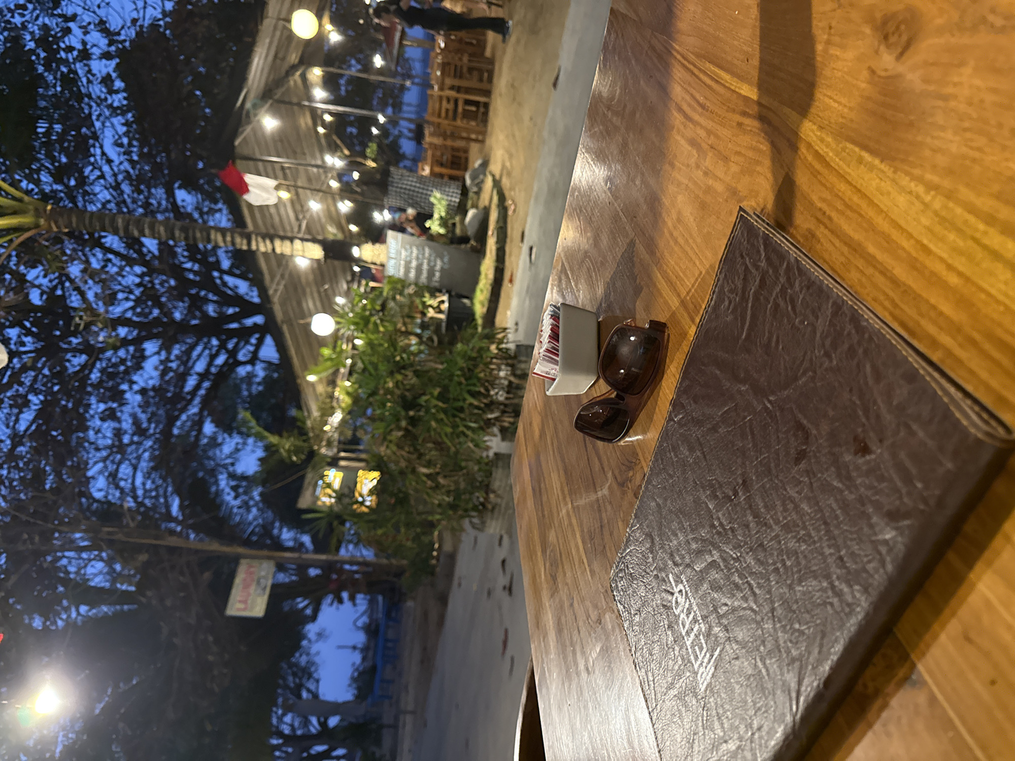 The image depicts a table at an outdoor restaurant in Denpasar, Indonesia, likely near a beach given the tropical foliage and the ambient lighting. In the foreground, a dark brown leather menu lies on a wooden table. To the right of the menu are a pair of brown sunglasses and a small white square holder filled with sugar packets and possibly straws. The background shows a covered seating area with numerous string lights and tables and chairs. Several trees, including a palm tree, are visible, with a red and white flag of Indonesia hanging from one. The sky is a deep blue, indicating it is evening or dusk. A sign that appears to say "LAUNDRY" is visible on a pole in the mid-ground. The overall atmosphere is relaxed and suggests a dining experience in a tropical location.