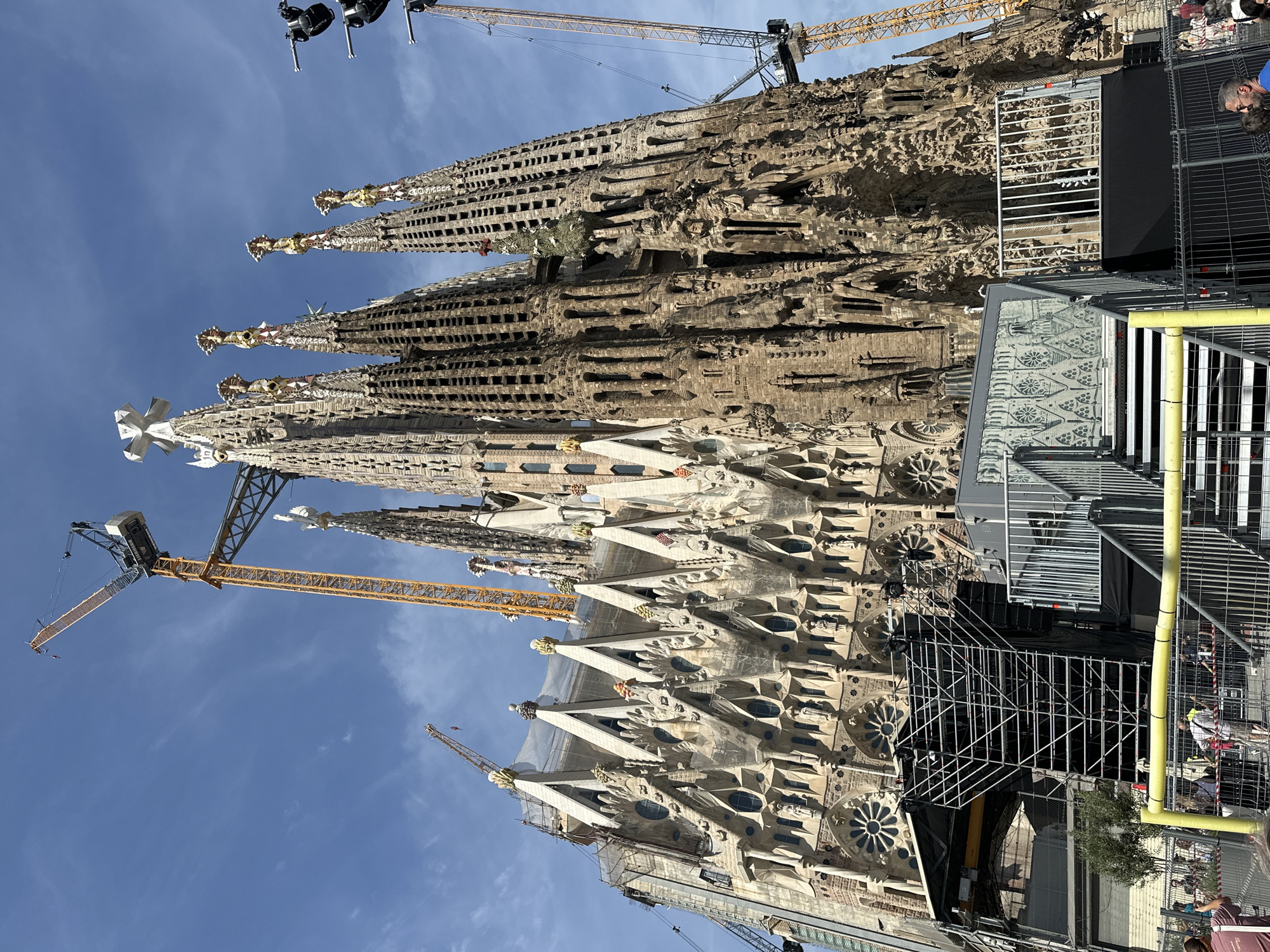 The image depicts the Sagrada Familia basilica in Barcelona, Spain, under construction on a clear day. The monumental structure, characterized by its intricate Gothic and Art Nouveau architectural elements, dominates the frame, showcasing numerous spires, detailed sculptural facades, and rose windows. Visible are at least three large tower cranes: a prominent yellow and grey crane extends high above the Passion facade on the right, another similar crane is positioned on the left, and a smaller, more distant crane is visible further left against the blue sky. Portions of the basilica, particularly on the left side, are covered with white netting and scaffolding, indicating ongoing construction or restoration work. In the foreground, a substantial temporary event structure is present, featuring black platforms, metal scaffolding, stairs with grey treads and white railings, and grey panels with decorative cutouts. Yellow metal barriers and additional metal fencing separate this structure from a small crowd of people. Individuals are visible in the lower left and right, standing behind the barriers, observing the scene. The sky is bright blue with scattered wispy clouds.