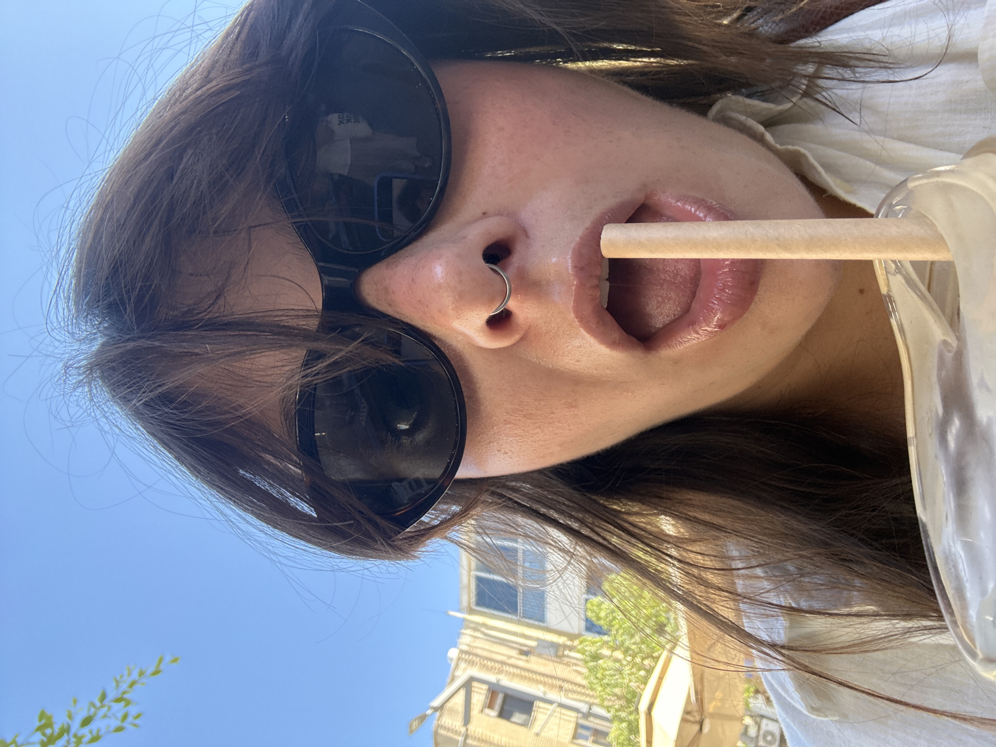 A woman is taking a selfie outdoors on a sunny day, holding a straw to her lips. She is wearing dark sunglasses and has a septum piercing. The background shows a clear blue sky, buildings, and some greenery, suggesting an urban environment. The lighting indicates it is daytime. The image captures a casual, relaxed moment.