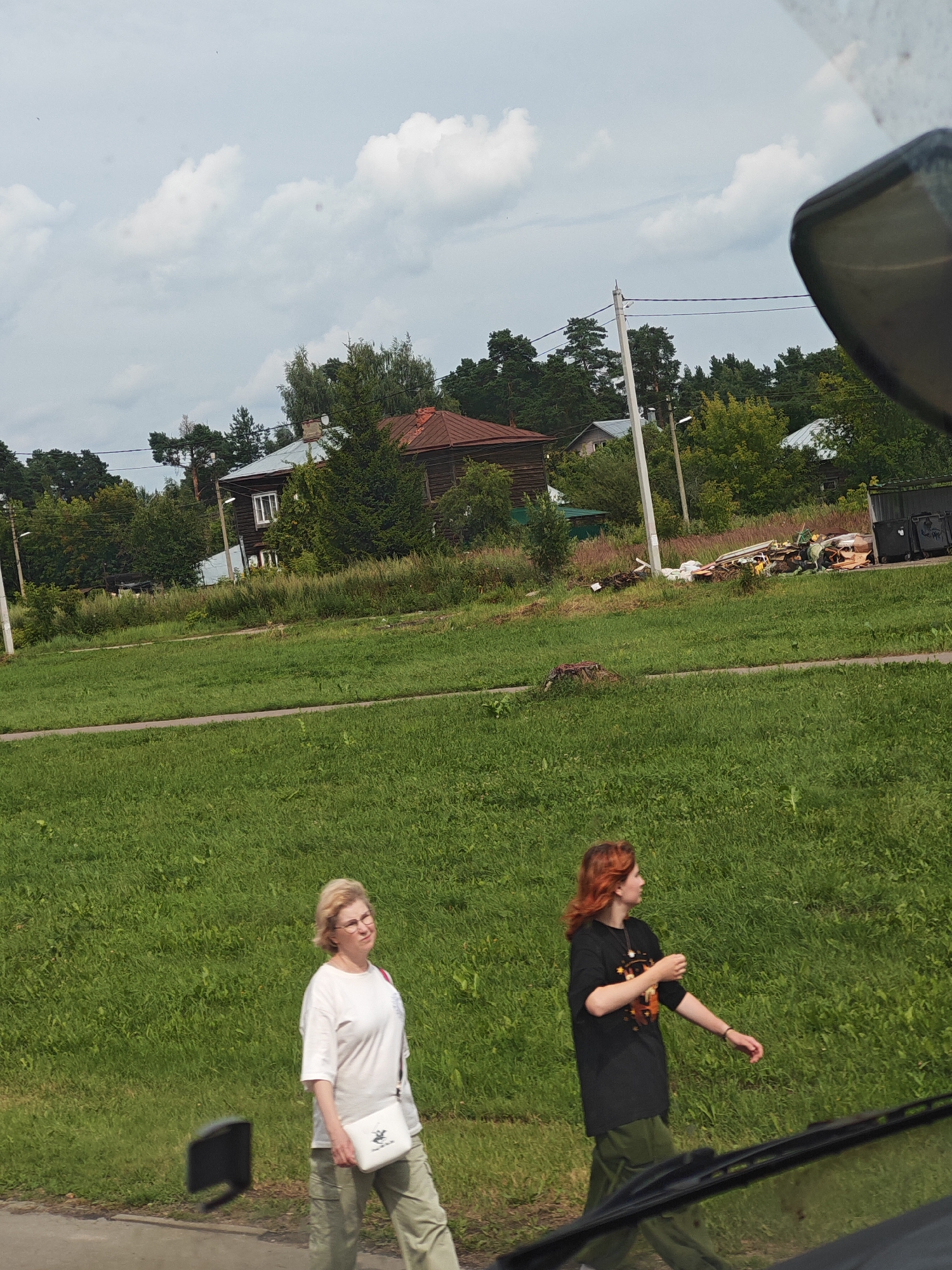 In the city of Likino-Dulyovo, Russia, on a cloudy day, two women are walking along a grassy area next to a paved path. The woman on the left, with blonde hair and glasses, wears a white t-shirt and light-colored cargo pants, carrying a white cross-body bag. The woman on the right, with red hair, is wearing a black t-shirt with a graphic design and dark green cargo pants. Behind them, a collection of discarded materials, including wood, plastic, and white bags, is piled near some dark trash receptacles. Further in the background, there are several houses, some with wooden exteriors and red roofs, surrounded by trees. Utility poles and power lines are visible. The overall environment is rural or suburban with abundant green vegetation.