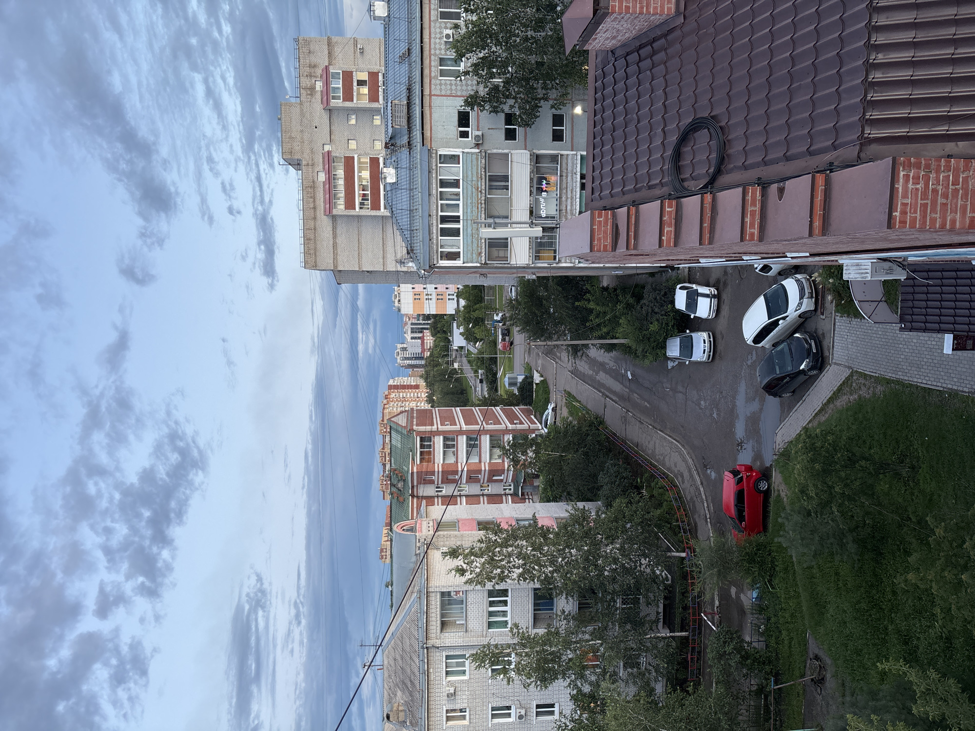The image depicts an aerial view of a residential area in Blagoveshchensk, Russia. The scene is dominated by apartment buildings of varying heights and architectural styles, constructed from brick and concrete. Power lines crisscross the sky between the buildings. In the foreground, a dark brown, tiled roof is visible. Below, a street winds through the neighborhood, with several cars parked along its side. These include a red SUV with its rear hatch open, a black sedan, and several white cars. Greenery, including trees and grass, is interspersed throughout the urban landscape, with some areas appearing somewhat unkempt. The sky is overcast with scattered clouds, suggesting either early morning or late evening. There are no visible people in the image.