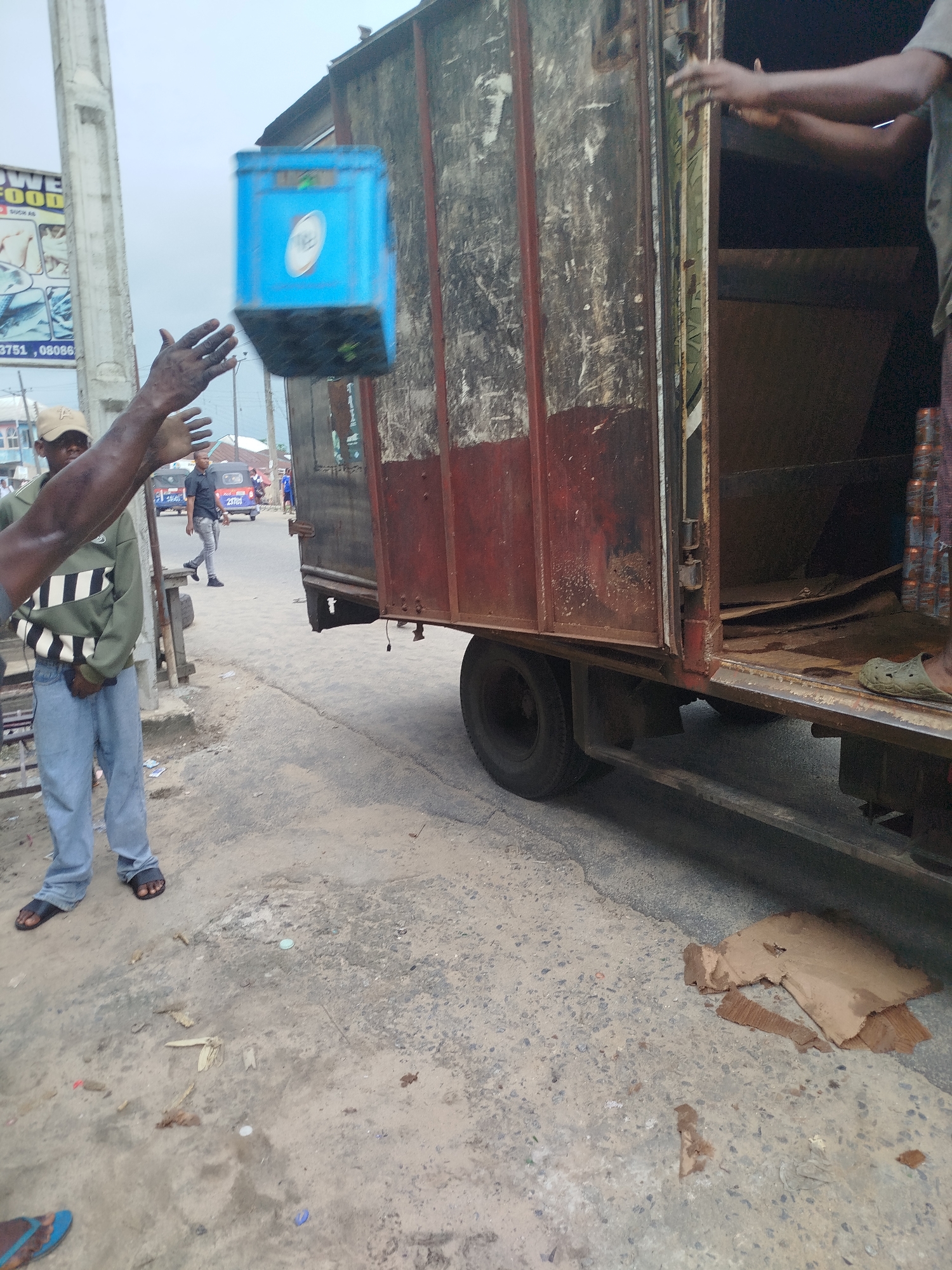 The media file captures an outdoor scene in Yenegoa, Nigeria. The primary subjects are two individuals, with a focus on one person actively engaged in throwing a blue container. A second person, partially visible and in the process of receiving or interacting with the thrown object, is also present. The scene is set on a dusty, uneven street beside a large, weathered truck. The truck appears to be a utility or cargo vehicle, with its side door open, revealing the interior filled with cardboard debris.
The activity depicted is the transfer of a blue container, possibly for goods or supplies, from one person to another. The person throwing the container has their arms extended, mid-throw, and the container is captured in mid-air. The other person's hands are also extended, suggesting they are about to catch or have just caught the container.
Notable details include the overcast sky, suggesting a cloudy day with diffused light. The environment appears to be an urban or semi-urban setting, given the presence of the road, the truck, and a pole with signage in the background. The signage has partial text visible, including "FOOD" and numbers like "3751, 08086", indicating a commercial area. The ground is littered with debris, including pieces of cardboard and plastic. The overall impression is one of practical, perhaps informal, labor taking place. The emotion conveyed is difficult to ascertain due to the image quality and focus, but the action suggests efficiency and coordination.