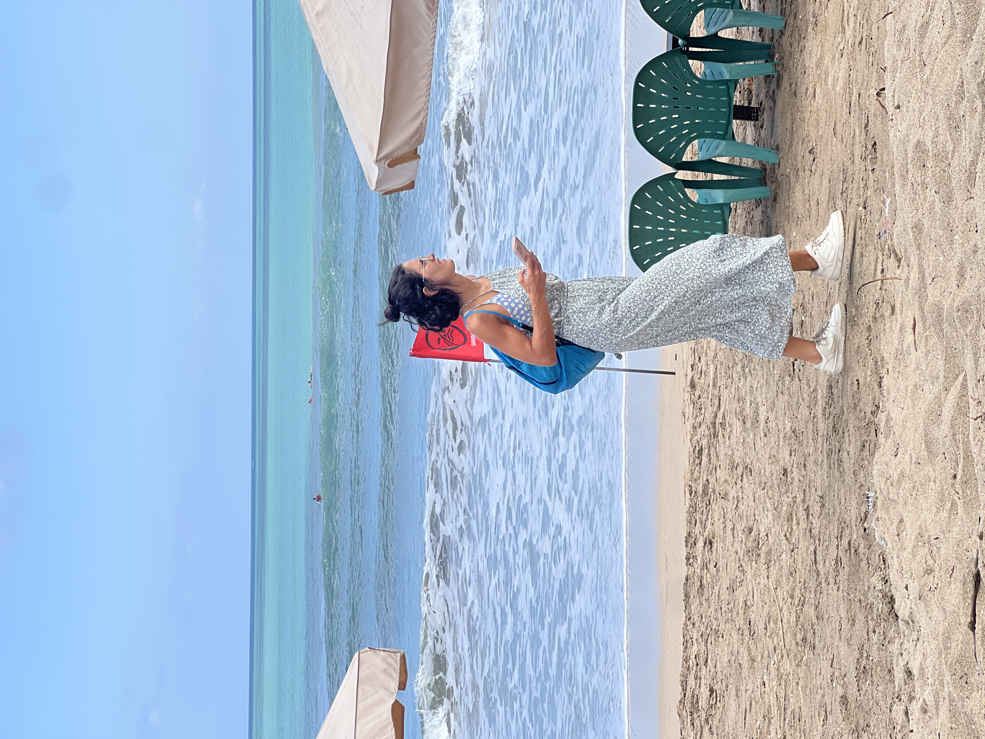 A woman with dark hair pulled into a bun, wearing glasses, a light blue patterned dress, and white sneakers, walks on a sandy beach in Kuta, Indonesia. She carries a bright blue backpack and is looking at her phone. In the background, the turquoise ocean meets the horizon under a clear blue sky. Waves are breaking on the shore, and several green plastic chairs are lined up on the sand. A red flag with a white symbol is planted in the sand behind the woman, and two beige beach umbrellas are visible, one on the left and one on the right side of the frame. A few surfers can be seen in the distance on the water.
