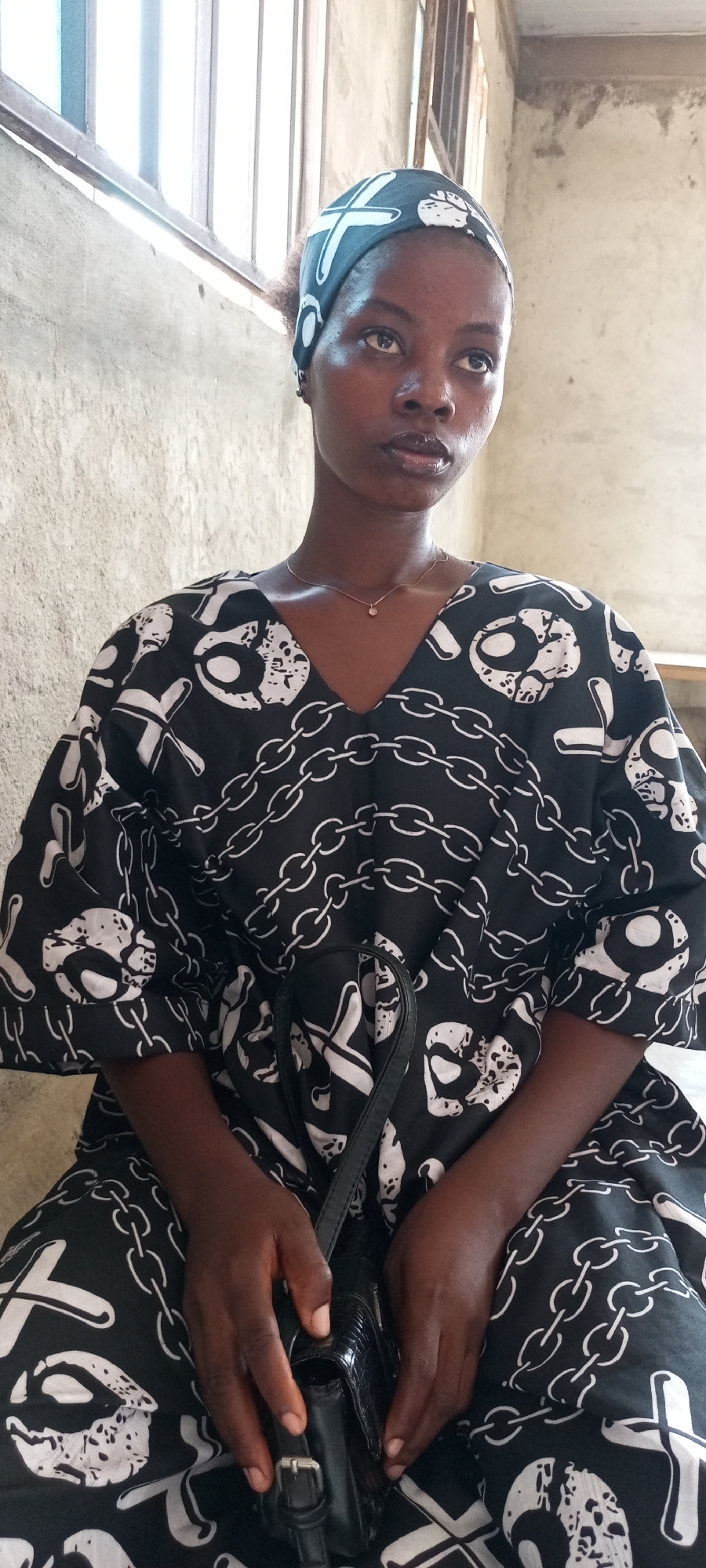 A medium shot depicts a woman seated indoors. She wears a black headscarf and a black and white patterned dress. A small black purse hangs from her shoulder, and her hands rest on it. Behind her is a textured wall and a barred window. The image appears to be taken in Lokoja, Nigeria.
