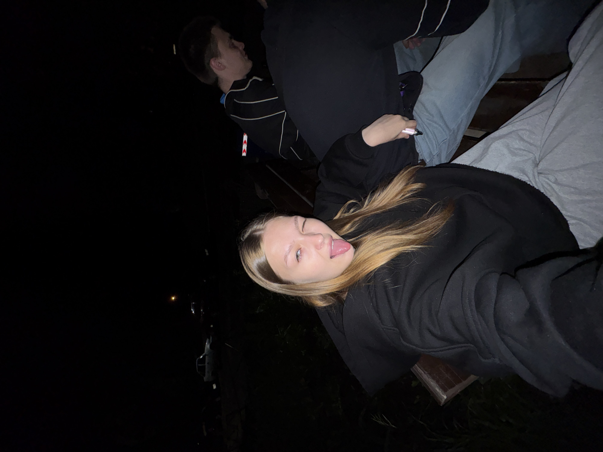 The image depicts an outdoor scene at night in Chaikovsky, Russia. Three individuals are present.
In the foreground, a young female with long, straight, light brown hair is seated, directly facing the camera. She is wearing a dark hooded sweatshirt and light gray trousers. Her right eye is closed in a wink, and her tongue is extended. Her left hand, positioned near her hip, holds a slender, light pink cylindrical object, possibly an electronic cigarette. She is seated on a dark wooden surface, likely a bench.
To her right and slightly behind, a young male with short, dark hair is also seated. He is wearing a dark track jacket with prominent white horizontal stripes across the chest and arms. His head is turned to the right, and his gaze is directed away from the camera. Behind him, a reflective red and white chevron pattern, characteristic of a road sign or barrier, is visible.
Between the female and the male subject, a third individual is partially visible, primarily their back and left arm. This person is wearing dark clothing, potentially a black hoodie, and light blue jeans. They are seated, with their left arm resting near the female's leg.
The environment is dimly lit, indicating nighttime. Sparse ambient light illuminates the subjects, possibly from an off-camera source or a flash. In the dark background, silhouettes of several vehicles are discernible, including an older model white sedan. A single, distant orange light source, consistent with a street lamp, is visible in the far background. Dark vegetation or ground cover outlines are present in the foreground and midground, suggesting a natural or unkempt outdoor area, possibly near a road or parking lot.