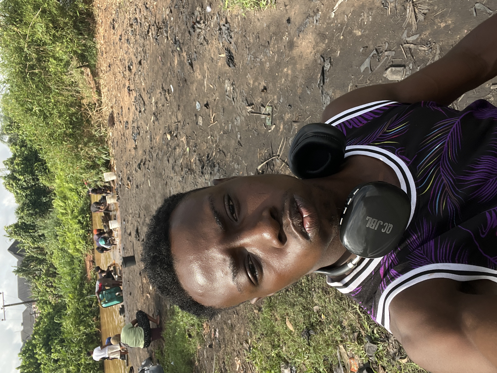 The photo shows a man wearing a black and purple jersey with white stripes and black JBL headphones around his neck. He is standing near a river or stream in Obosi, Nigeria. Several people are washing items in the water. The ground surrounding the area is muddy and littered with debris. There is also vegetation alongside the water.