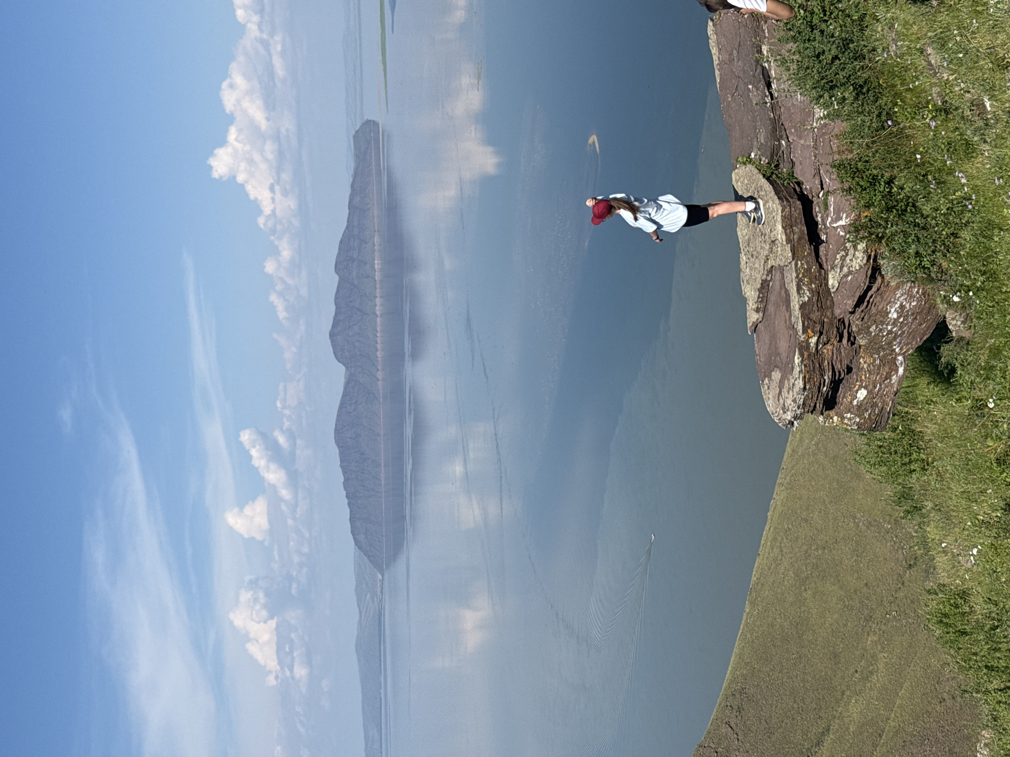 A person stands on a rocky precipice overlooking a vast, calm body of water. The person, seen from behind, wears a light blue top, black shorts, and a red cap. They are facing away from the camera, looking out at the expansive vista of the water and a large, dark, mountainous island in the distance. The sky is blue with scattered white clouds, reflecting on the water's surface, which also shows the wake of a distant boat. The foreground is dominated by green, grassy slopes and the jagged, reddish-brown rocks of the overlook. Another person's arm is visible on the far right edge of the frame. The location is a scenic overlook in Ust'-Abakan, Russia.