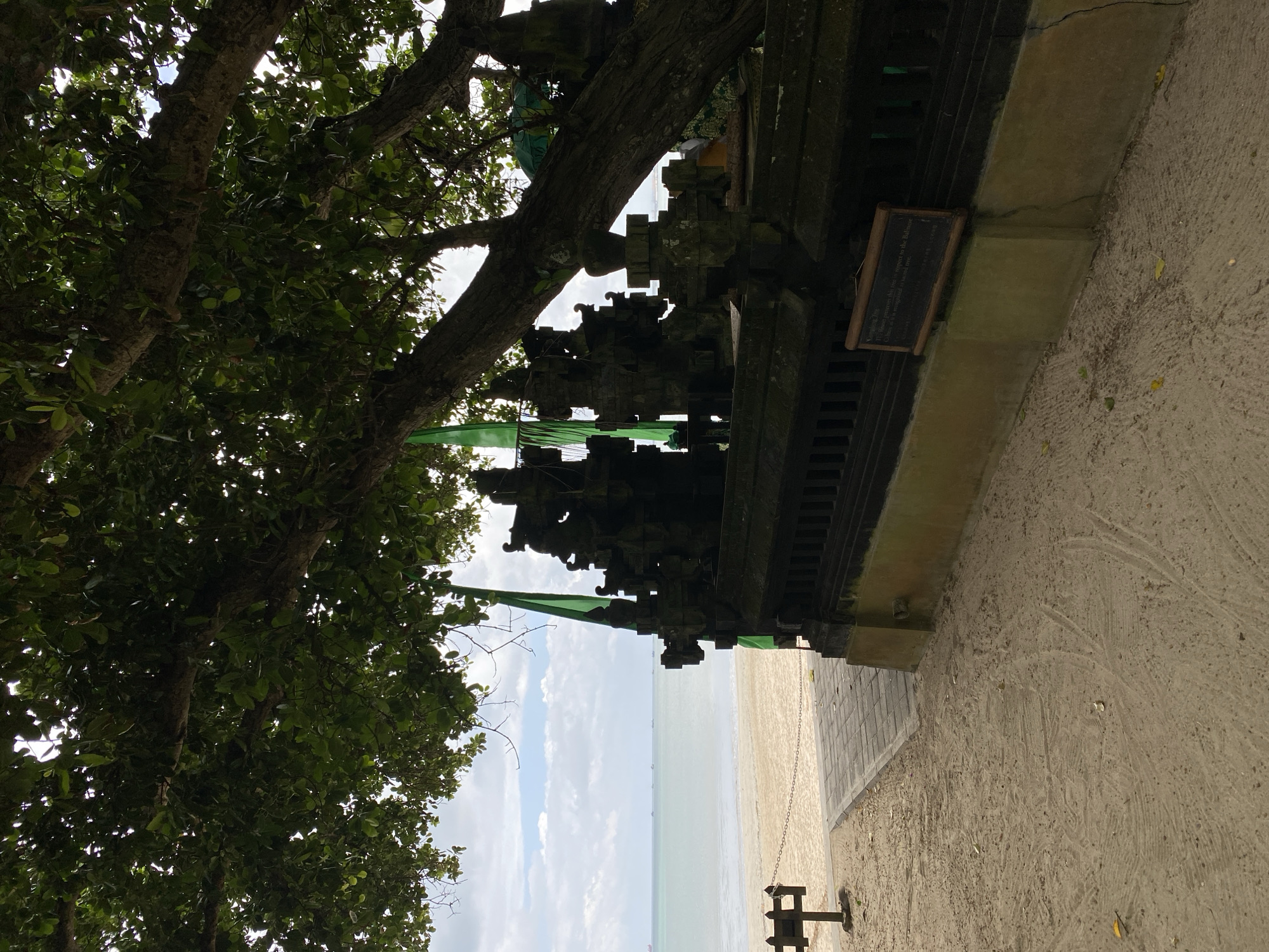 This is an outdoor, eye-level shot, featuring part of a Balinese temple in Kuta Selatan, Indonesia. The temple is made of gray stone and appears to be situated next to a beach. Some green flags adorn the temple. A sign hangs on the side of the temple. A tree with abundant green foliage partially obscures the temple. The sky is visible in the background, displaying a mix of clouds and blue patches. The ground adjacent to the temple is sandy.
