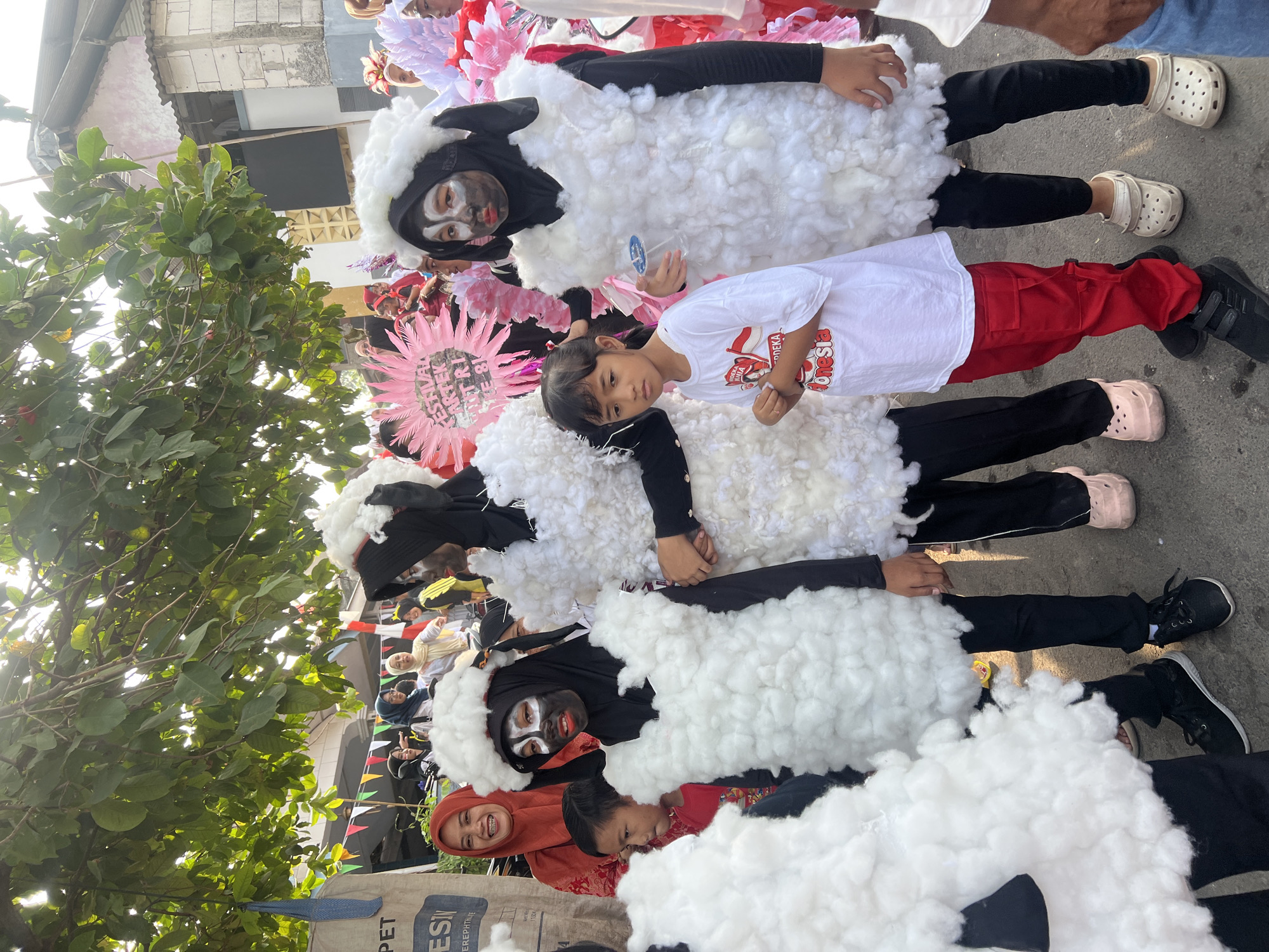 In Klapanunggal, Indonesia, three children, two wearing sheep costumes made of white fluff over black hijabs and pants, stand in a street. The child on the left has black face paint with white accents around the eyes and mouth, and red lipstick. The child in the center has black face paint with white accents around the eyes and mouth. The child on the right also has black face paint with white accents around the eyes and mouth. A young girl in a white t-shirt with red trim and red pants stands between the two sheep-costumed children. The t-shirt has an Indonesian flag design and "MERDEKA" and "INDONESIA" text. Another child in a red shirt and black pants is visible to the left. Behind them, adults and other children are present, some wearing colorful costumes. A large, pink, fan-like decoration with text is visible to the right of the center child. Bunting flags are strung across the background. The ground is paved.