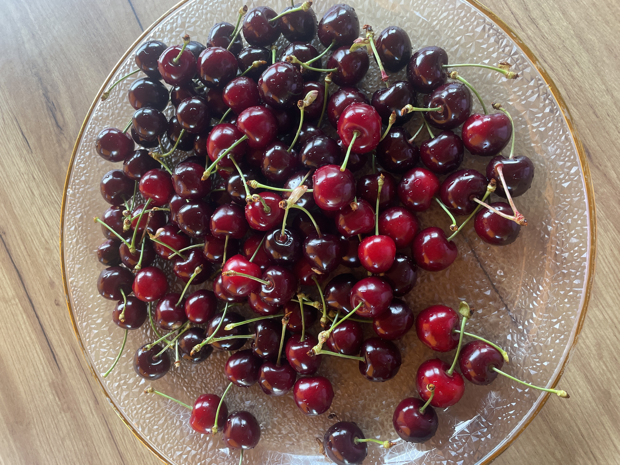 A transparent, textured glass plate with a gold-colored rim contains a substantial quantity of fresh cherries. The cherries, predominantly dark red to deep maroon, exhibit varying shades with some brighter red examples interspersed. Numerous small water droplets are visible on the skin of many cherries, indicating recent washing or freshness. Most cherries retain intact green stems, some individually attached, others in small clusters. The plate, filled mostly with the cherries, rests on a light to medium brown wooden surface, displaying discernible wood grain patterns. The illumination suggests natural light, creating soft reflections on the fruit and glass.