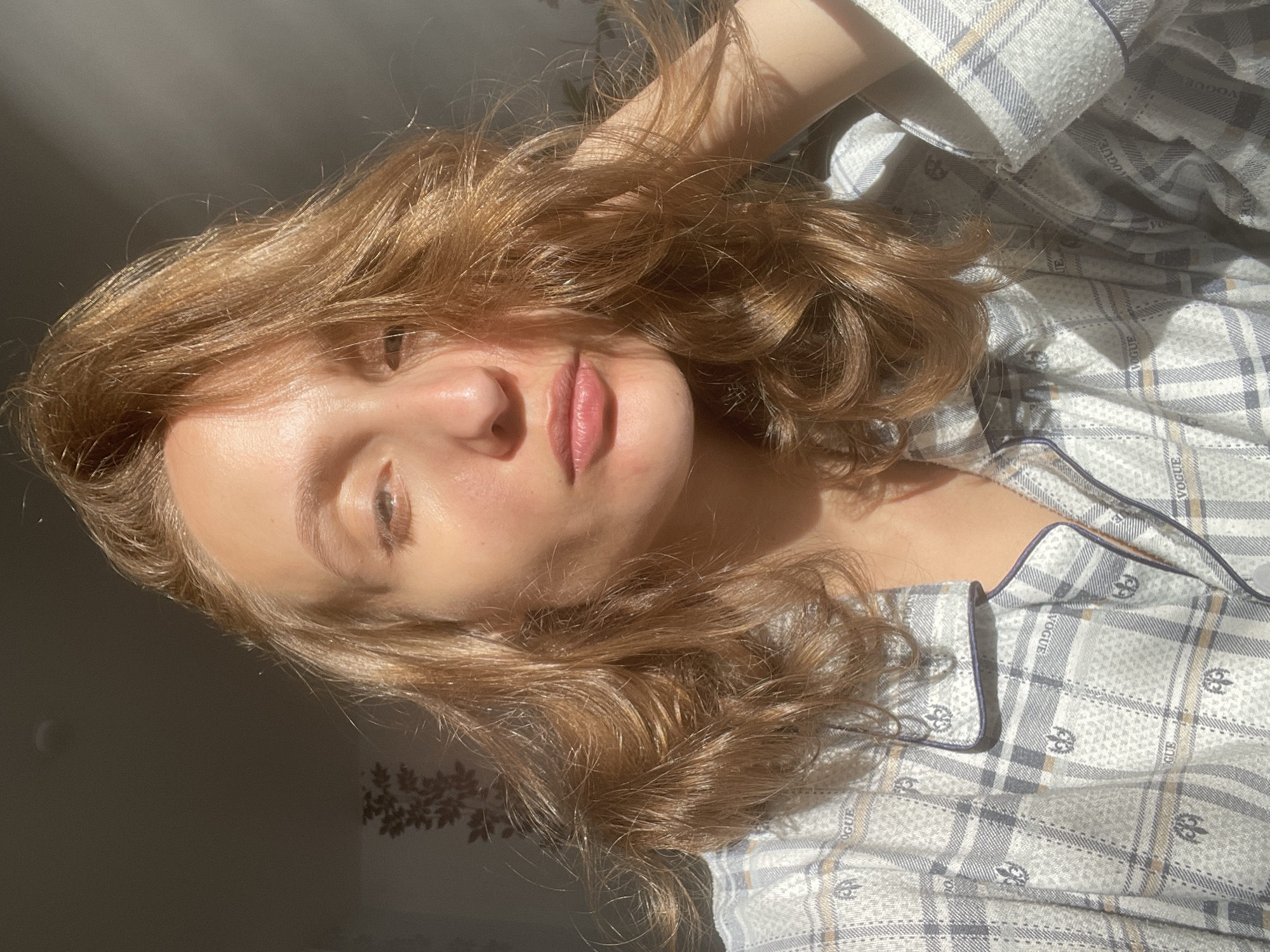 The media file is a close-up, high-angle shot of a young woman lying down, with her head resting on her arm. The lighting suggests it is daytime, with sunlight casting shadows and illuminating her face, which is angled slightly upwards. The woman has light brown, wavy hair that falls around her face. She is wearing pajamas with a plaid pattern in shades of grey and white, featuring a repeated "VOGUE" text and fleur-de-lis emblems. Her lips are slightly parted and painted with a subtle pink lipstick. Her eyes are partially closed, and her expression appears relaxed and perhaps sleepy. The background is dark, with a faint floral pattern visible on the wall behind her. The overall mood is intimate and tranquil, suggesting a private moment.