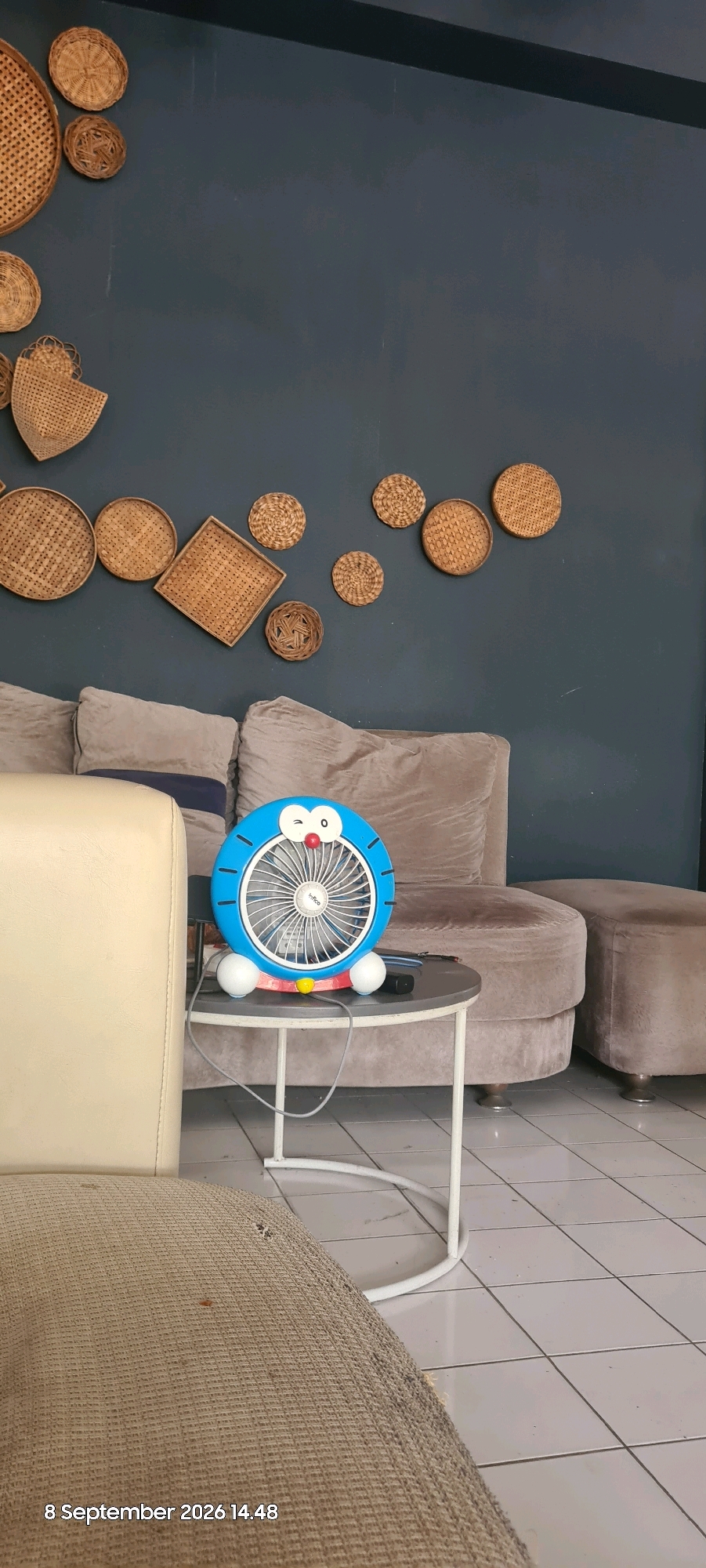 A Doraemon-themed fan sits on a small round table in front of a plush sofa in a room with a dark blue wall. The fan is blue with white spheres for feet, and has Doraemon's face with a red nose and a wink. The wall is decorated with several woven rattan circular and square wall hangings of various sizes, arranged artfully. To the left of the fan, a cream-colored armchair is partially visible. The room appears to be a living room or lounge area. The timestamp "8 September 2026 14.48" is visible in the bottom left corner, indicating the image was taken on September 8, 2026, at 2:48 PM.