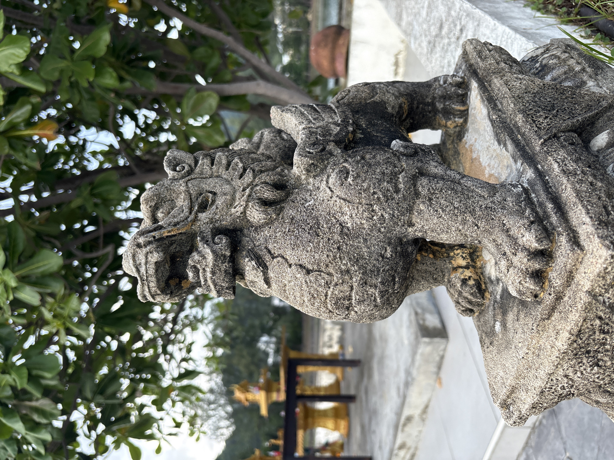 A stone statue of a mythical creature, possibly a Foo dog or lion, is the central subject of this image, located outdoors. The statue is weathered and gray, with visible texture and some gold discoloration on its paws. It is positioned on a stone ledge in the foreground, which appears to be part of a larger stone structure. Behind the statue, out of focus, are green trees with dappled sunlight filtering through their leaves. Further in the background, a low table with golden ornate stands and structures is visible, suggesting a shrine or altar. The ground appears to be paved with concrete. The overall setting suggests a location in a tropical or subtropical environment, possibly a temple or a garden area. The specific location is identified as Karon, Thailand. There are no people visible in the image.